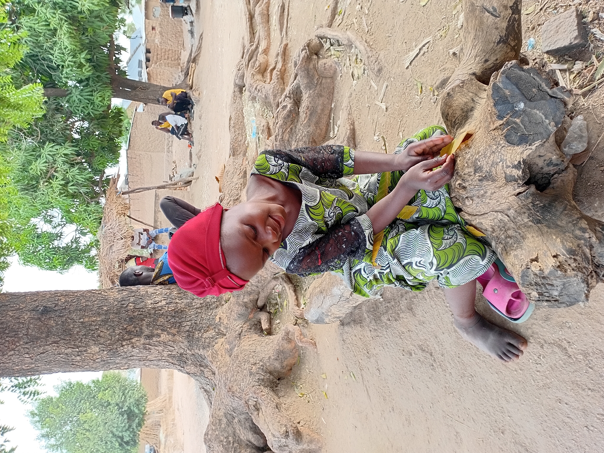 A young girl sits cross-legged on a weathered tree stump in Nata-Ala, Nigeria. She wears a vibrant green and white patterned dress with black lace sleeves and a red head covering. She holds a small yellow leaf in her hands. Her right foot is bare, while a pink sandal adorns her left. The stump is surrounded by sandy ground, and a large tree trunk extends upwards on the left side of the frame. In the background, a group of people gather near buildings under the shade of a tree.