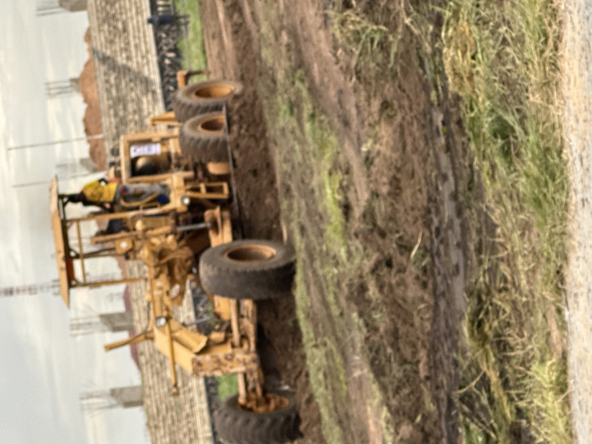 A yellow grader is operating on a dirt surface, likely a construction site. The grader is a heavy machine with large wheels and a blade used for leveling ground. The operator, wearing a yellow shirt, is visible in the cab of the grader. In the background, there are unfinished concrete structures, possibly part of a stadium or large building. The terrain is uneven and appears to be under development. The overall scene suggests construction activity is in progress.