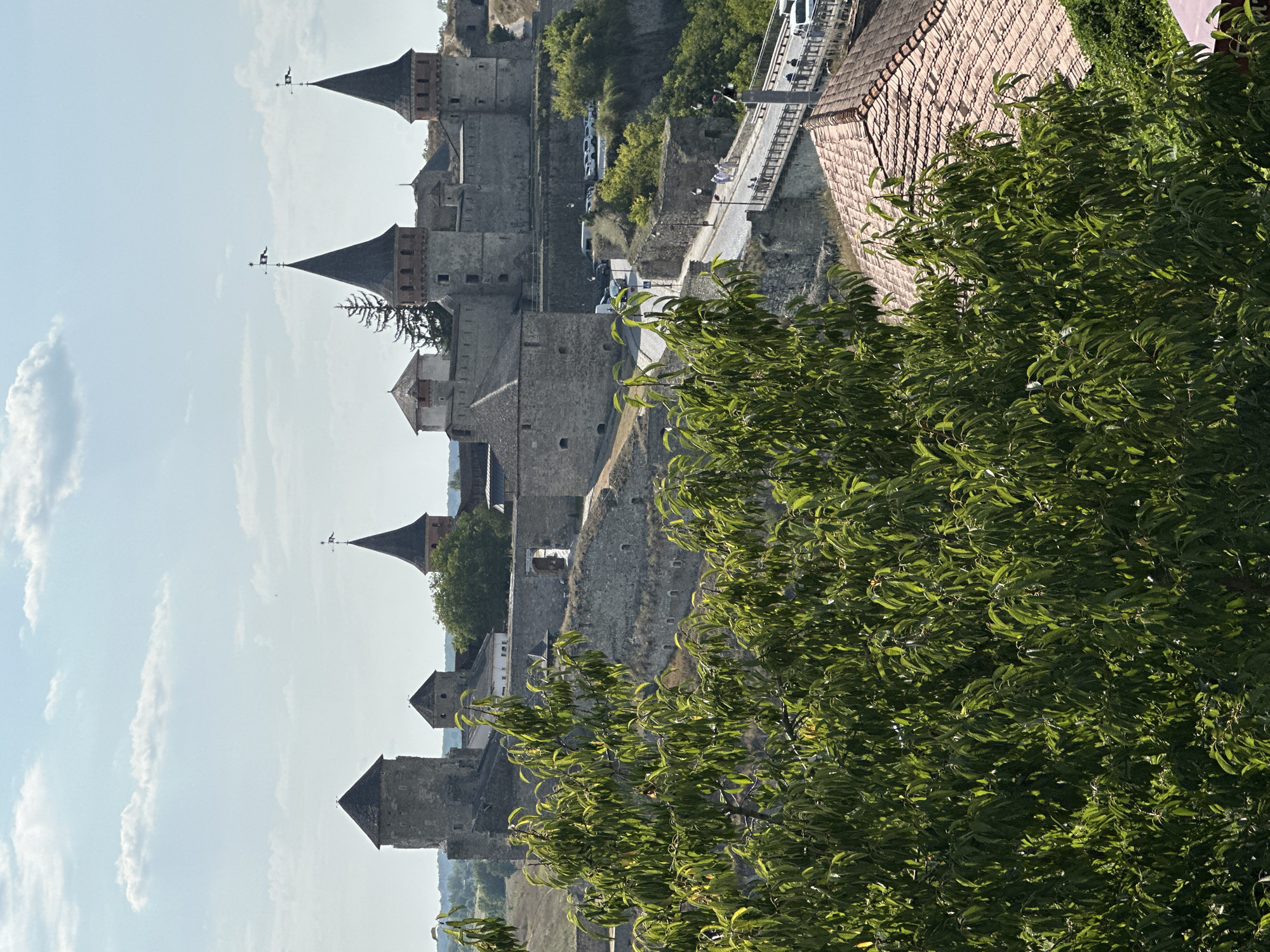 A distant, high-angle, full shot captures the historic Kamianets-Podilskyi Castle in Ukraine under a bright, lightly clouded sky. The stone fortress, with its various towers, some capped with dark, conical roofs and topped with flags, sprawls across a rocky, elevated landscape. In the foreground, vibrant green foliage from trees partially obscures the view, adding a natural element to the scene. A bridge with railings and pedestrian traffic spans a lower level, with cars visible on a road winding towards the castle. The architecture suggests a medieval or Renaissance era, with weathered stone walls and prominent defensive structures. The overall impression is one of a well-preserved historical site situated within a picturesque, rugged environment.
