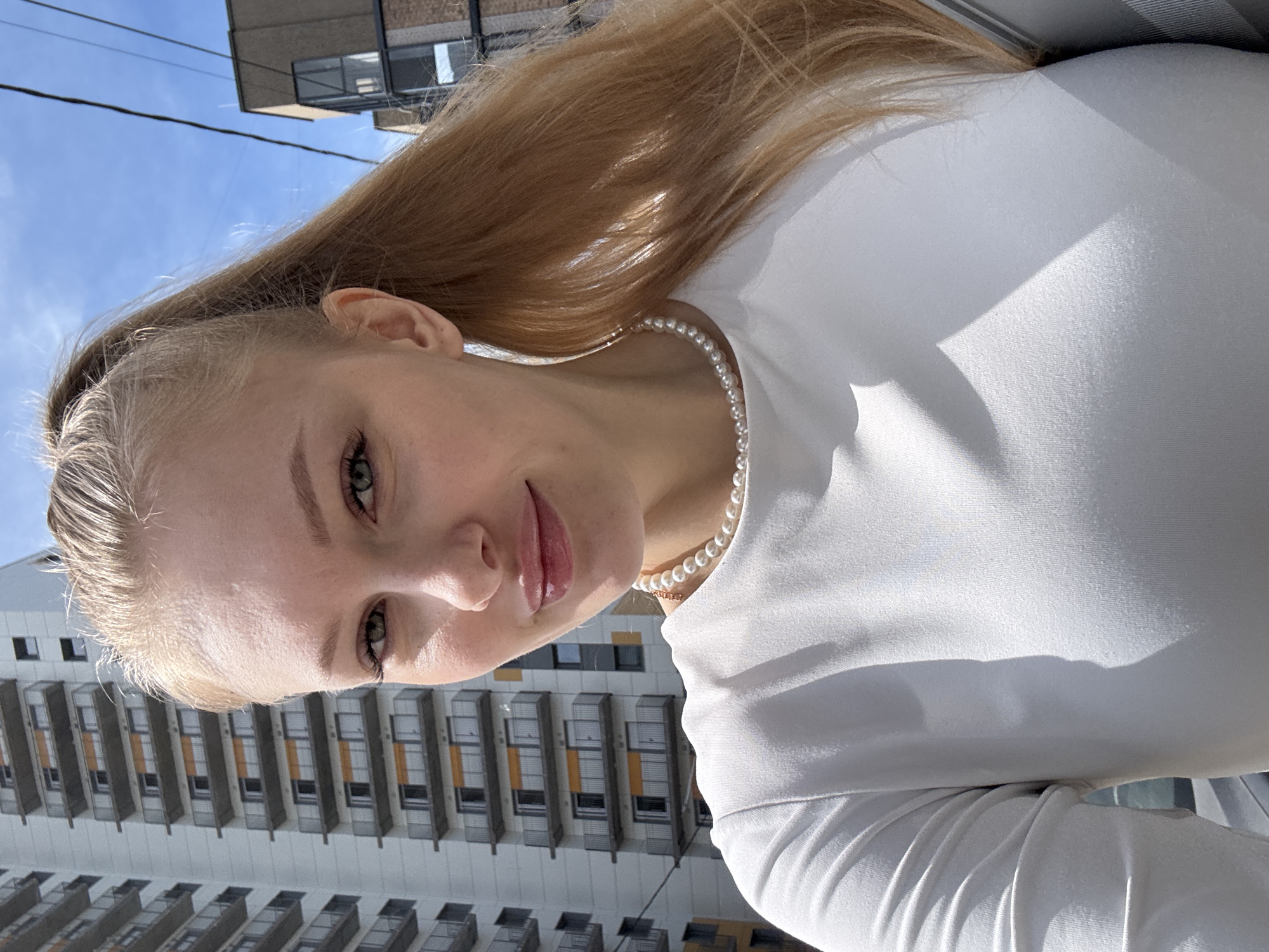 A young woman is featured in a brightly lit portrait, captured against an urban backdrop on a sunny day. She is seen from the chest up, with her head tilted slightly, looking directly at the camera with a subtle, serene smile. Her long, light blonde hair, appearing golden in the strong sunlight, flows over her shoulders and back. She has fair skin, light-colored eyes (likely green or hazel), and natural-looking makeup with slightly glossy lips. Around her neck, she wears a delicate single-strand pearl necklace, complementing her simple, close-fitting white long-sleeved top.
The background showcases modern residential buildings under a clear, vibrant blue sky. A tall, multi-storied building with numerous balconies and windows is prominently visible to the lower left, exhibiting contemporary architectural design common in urban settings like Perm, Russia. The direct sunlight creates bright highlights on the woman's hair and clothing, while also casting soft shadows that add dimension to the buildings and her attire. The overall atmosphere is one of brightness and freshness, indicative of pleasant, clear weather. No visible text is present in the image.
