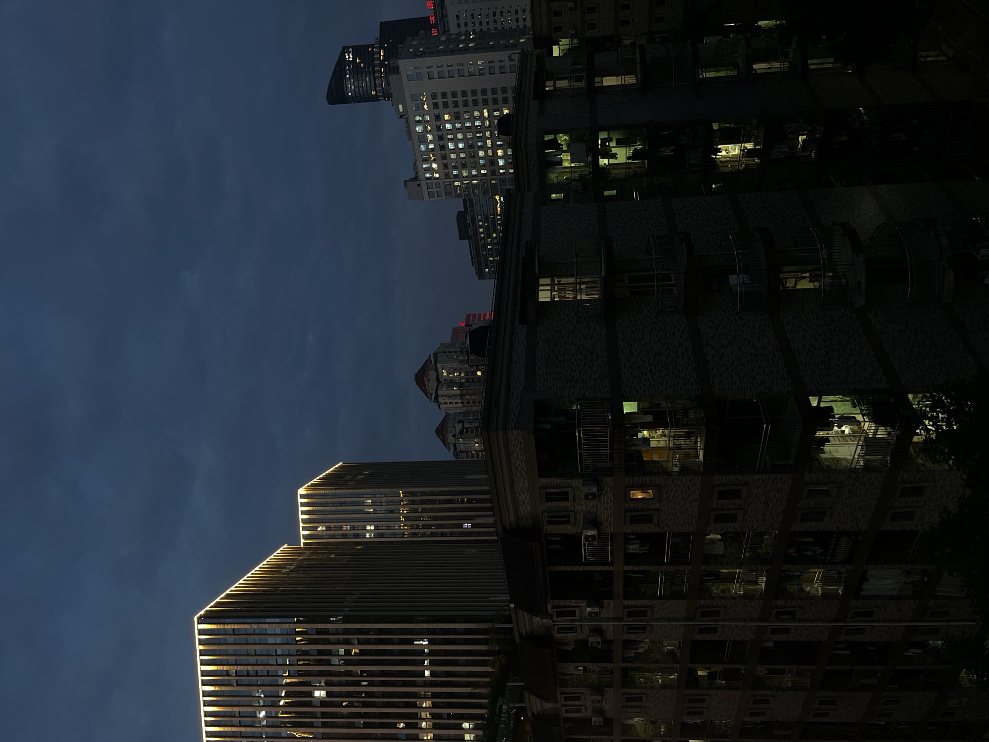The media file presents an urban night scene in Yuzhong District, China, showcasing a dense cluster of high-rise buildings under a dark blue-grey sky. The image is oriented vertically, showing the towering structures reaching towards the top of the frame.
On the left, a prominent modern skyscraper features a sleek design with horizontal bands and striking golden architectural lighting illuminating its edges and upper sections. Many windows on this building reflect the dim ambient light, and some interior lights are visible.
To the right, a multi-story building with a textured facade, possibly brick or stone, stands out. This structure has numerous windows, many of which are brightly lit, revealing glimpses of interiors, possibly offices or residential spaces, with what appear to be furniture or partitions. Several windows or balconies on this building are fitted with dark grilles or protective cages, and some also have visible air conditioning units.
In the mid-ground and background, other tall buildings are visible, some appearing older in design, others modern. Many of these buildings display a grid pattern of small, illuminated windows, suggesting active occupancy. On one of these mid-ground buildings, a red sign with visible Chinese characters can be discerned; it appears to spell "交通银行" (Jiāotōng Yínháng), translating to "Bank of Communications."
The overall scene captures the quiet grandeur of a major city at night, with no discernible human activity but abundant evidence of urban life through the myriad illuminated windows. The clear, dark sky and the contrast between the dark building masses and their lit windows create a serene yet imposing atmosphere.