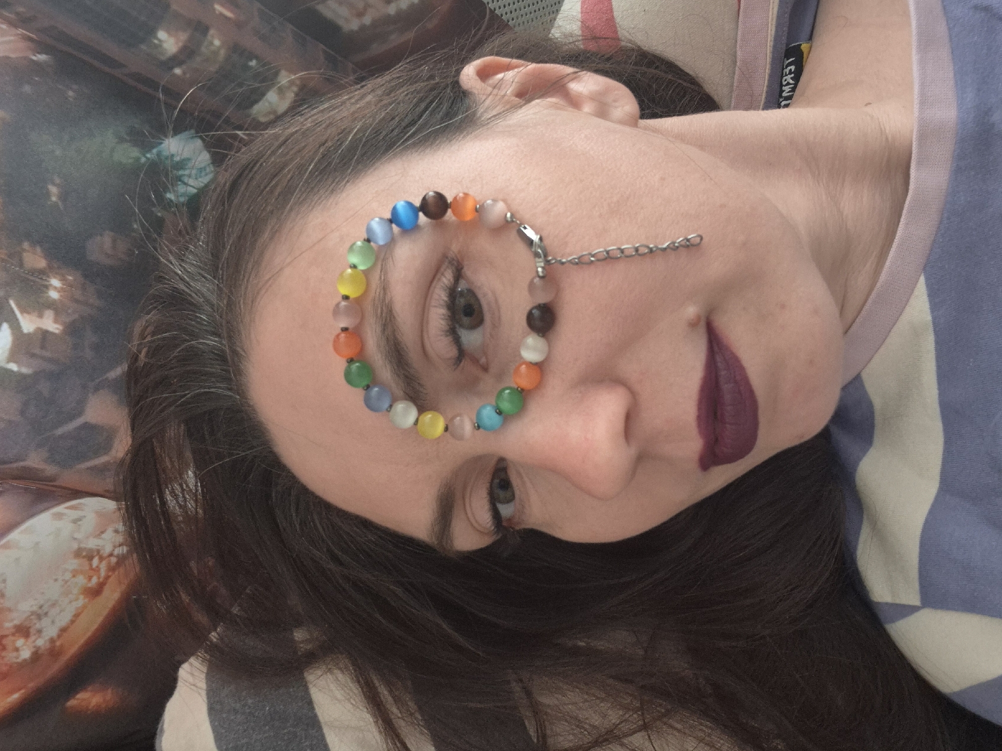 The image shows a woman with dark hair lying down, wearing a colorful beaded bracelet around her eye, connected by a chain to her cheek. She has dark lipstick on. The background consists of a pillow or bedding with a textured pattern and what appears to be part of a cityscape image. The specific location within any city is unknown.
