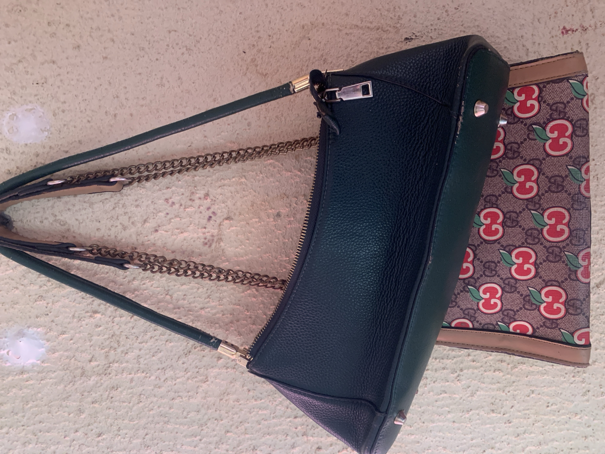 The media file is a close-up, overhead shot of two bags resting on a textured, light-colored surface, possibly a wall or floor. The main subject is a dark green, pebbled leather handbag with a gold zipper and chain strap. The bag is positioned diagonally, with its top facing upwards. A second bag, a flat wallet or clutch, is partially visible underneath the handbag. This second item features a repeating pattern of the Gucci logo intertwined with red cherry illustrations.
Notable details include the texture of the surfaces and the materials of the bags. The leather of the handbag appears somewhat worn, with a few scuffs visible, particularly near the strap attachment. The metal hardware on the handbag, including the zipper pull and strap connectors, is gold-toned. The chain strap is a thick, interlocking link style. The Gucci patterned item has a beige background with the brown interlocking "G" logo and vibrant red cherries with green leaves. The edges of this item have a thin, tan leather trim.
No people are visible in the image, and there is no indication of the time of day or weather. The setting is ambiguous due to the close-up nature of the shot, but the textured surface might suggest an indoor environment. There is no discernible text beyond the Gucci branding on the patterned item. The overall impression is a still life arrangement of fashion accessories.