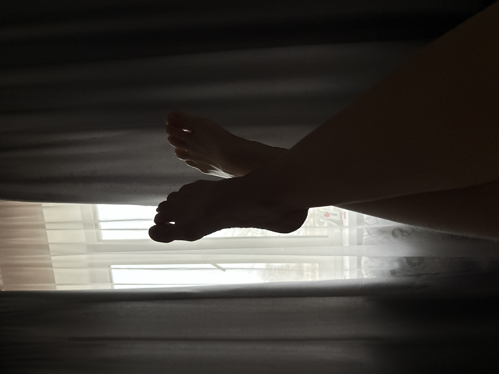 The image shows a view of two feet and legs through a narrow opening between dark, heavy curtains or blinds. Behind the feet, light filters through sheer white window curtains, suggesting a window. The scene is dimly lit. There is no information available in the image to determine the location of the image in the city of Chaikovsky, Russia. The only details present are feet, legs, curtains, a window, and general lighting conditions.