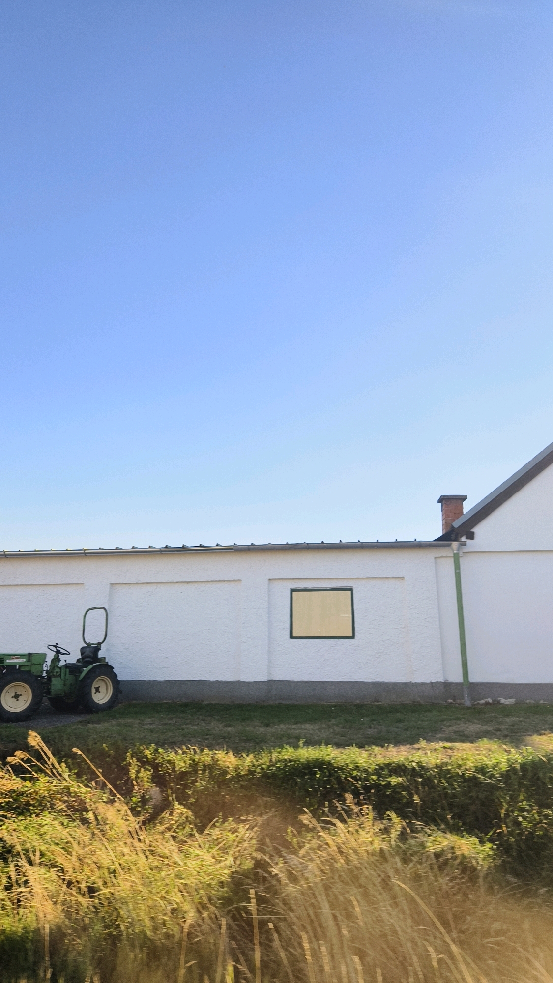 The image depicts an outdoor scene under a clear blue sky, likely taken in Fels am Wagram, Austria. A white building with a dark-tiled roof and a brick chimney dominates the right side of the frame. The building has a single square window with a yellow pane. To the left of the building, a small green tractor is parked on a strip of grass. In the foreground, tall, dry grasses and vegetation are illuminated by sunlight, creating a blurred effect. The overall scene suggests a rural or agricultural setting on a bright day.