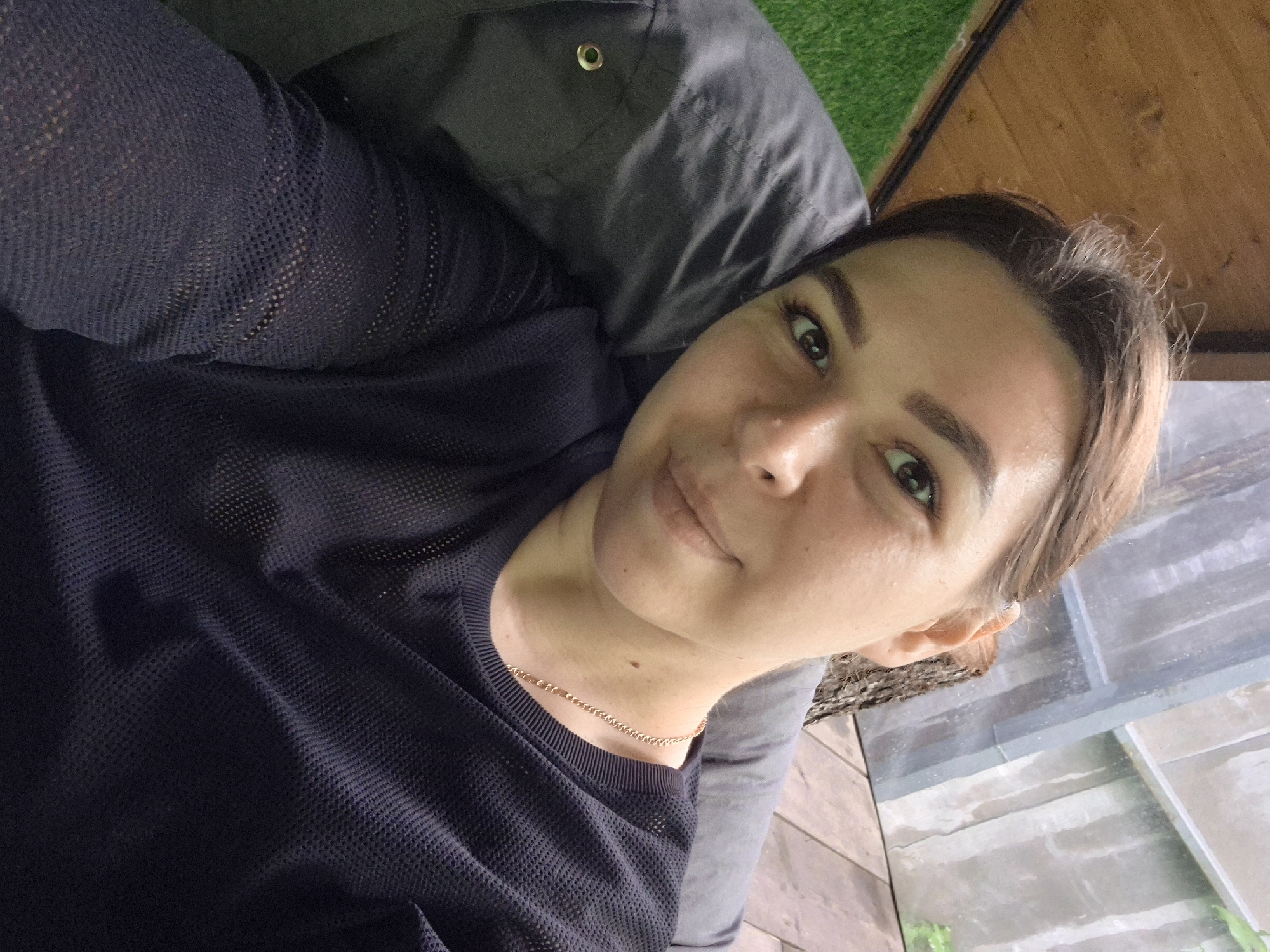 A selfie of a woman with brown hair pulled back is taken outdoors. She is wearing a dark blue mesh shirt and a delicate gold chain necklace. Behind her, a gray cushion is visible, as well as wooden planks and artificial turf. The background suggests an outdoor seating area or patio, possibly in a park or backyard setting. The lighting indicates it is daytime. The woman is looking directly at the camera with a slight smile. The location is identified as Izhevsk, Russia.