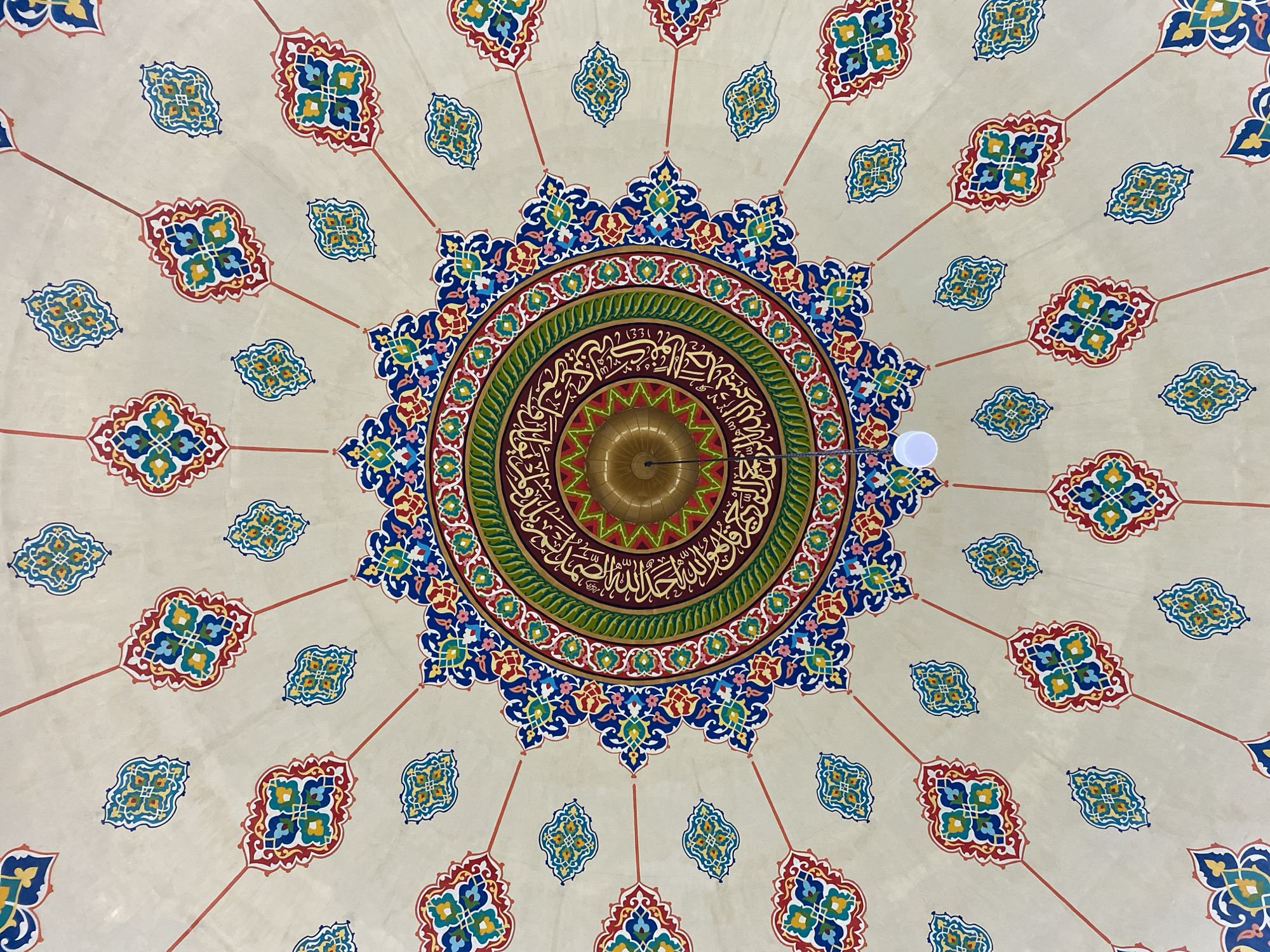 The image shows the interior dome of a mosque with intricate geometric and calligraphic patterns painted in vibrant colors. The central medallion features Arabic calligraphy surrounded by concentric rings of floral motifs in blue, red, green, and gold. Radiating outwards from the center are red lines connecting to smaller, repeating decorative elements in similar colors. The dome is a pale beige color, serving as a canvas for the elaborate artwork. A single light fixture hangs from the center, and another smaller light bulb is visible near the upper edge of the frame. The artwork suggests a traditional Islamic architectural style. Without further information, the specific location within Lhokseumawe, Indonesia, cannot be determined solely from the visual content of the image.