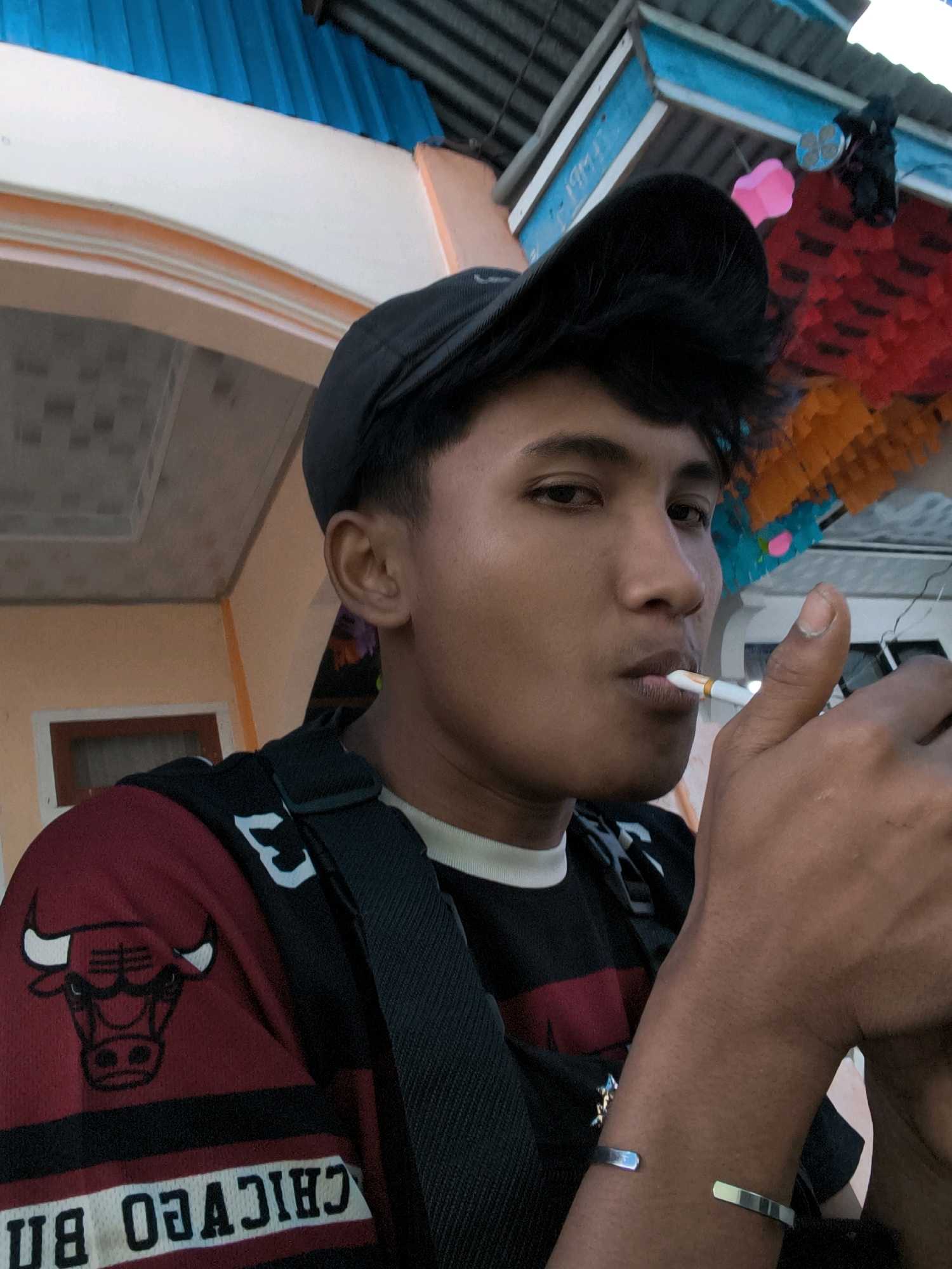 A young man, wearing a black baseball cap and a maroon and black Chicago Bulls jersey, holds a cigarette to his mouth and lights it. He is wearing a silver bracelet on his left wrist. He is positioned outdoors, with a building in the background that appears to be in a residential area. The building has a blue corrugated metal roof and a pale orange and cream-colored facade. Colorful, layered paper decorations are visible on the right side of the frame, suggesting a festive or cultural event may be taking place. The man's gaze is directed towards the camera. The image was captured in Sungai Penuh, Indonesia.