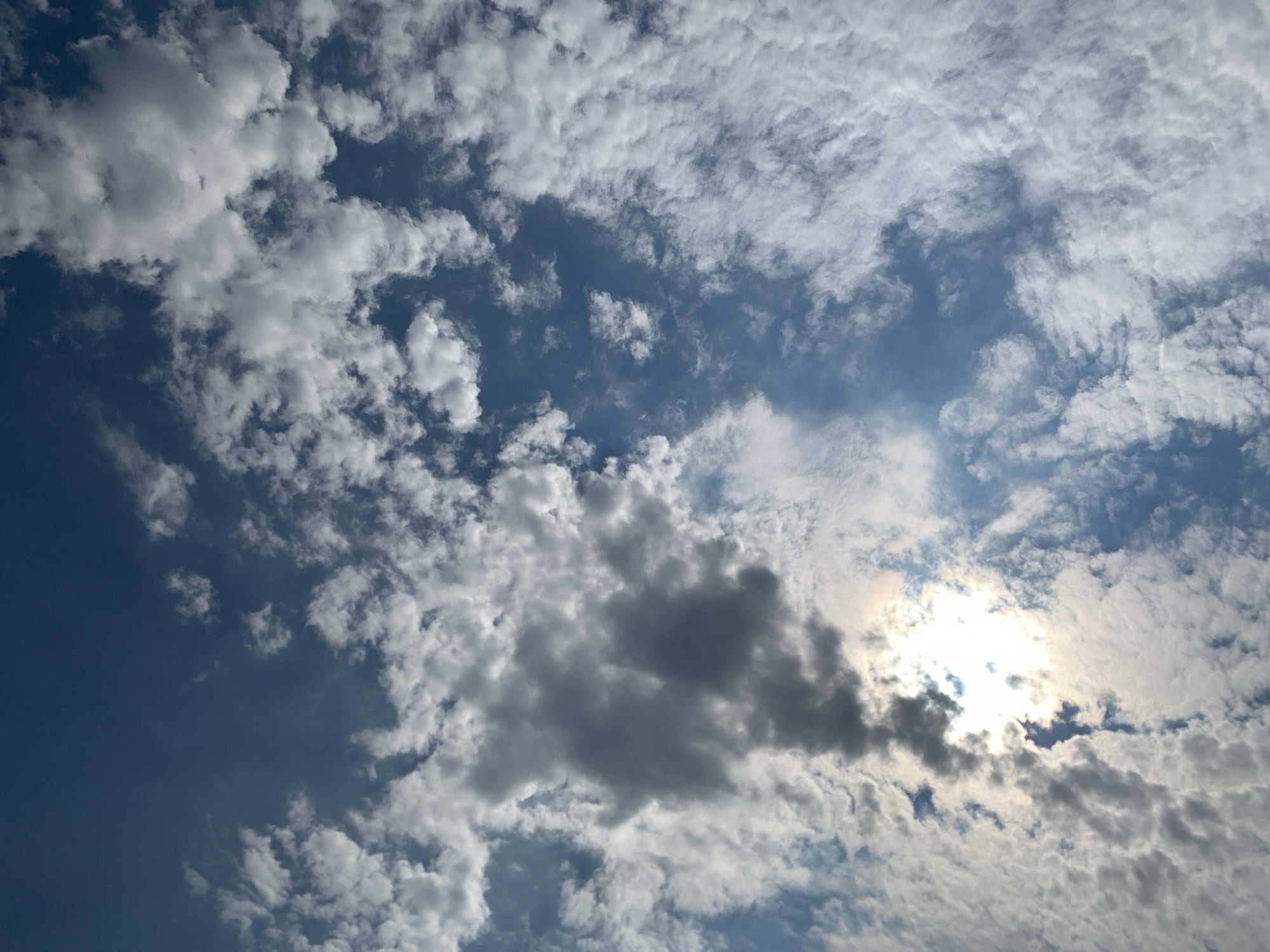 A wide-angle shot captures a dynamic sky filled with a mix of white and dark gray clouds against a deep blue backdrop. The sun is partially obscured by clouds, creating areas of bright illumination and shadowed contrast within the sky. The composition is entirely focused on the atmospheric elements, with no earthly objects or human presence visible. The scene is devoid of any specific landmarks that would indicate a location within Khazar Raion, Azerbaijan.