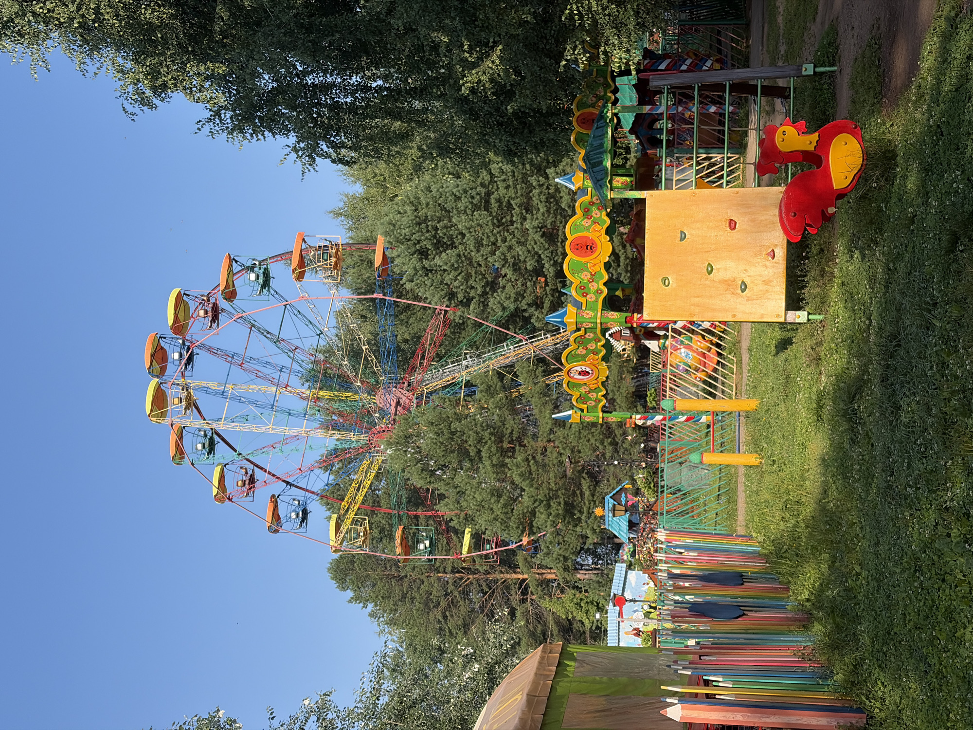 The image depicts a colorful amusement park situated in Chaikovsky, Russia. In the background, a large Ferris wheel with brightly colored cabins is prominently displayed against a clear blue sky, surrounded by lush green trees. In the foreground, a playground area is visible, featuring a climbing wall, a spring rider shaped like a rooster, and a fence constructed from multicolored wooden posts resembling pencils. Several other playground structures and rides, adorned with vibrant decorations, are also present. The scene is bathed in sunlight, casting shadows on the grassy ground. No people are visible in the image.