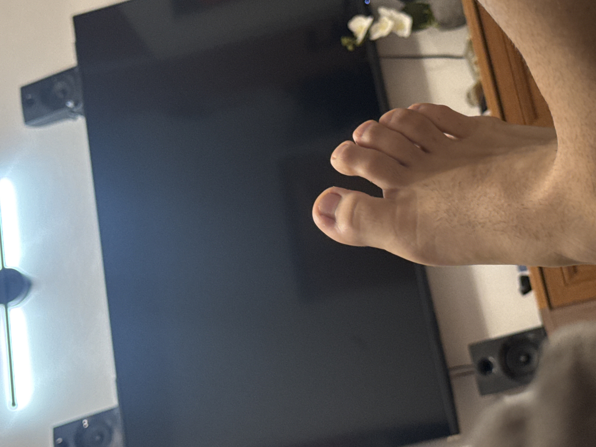 An interior photograph, taken from a low angle, depicts a person's bare foot in the immediate foreground, positioned in what appears to be a relaxed, reclined posture. The foot is light-skinned with visible hair on the instep and toes, which are slightly curled. The background features a large, black, flat-screen television prominently centered, likely turned off or displaying a dark image. Above the television, two compact black speakers are situated on either side of its top edge. A fluorescent light fixture, illuminating brightly, is visible on the white ceiling directly above the television, with a dark circular fixture positioned centrally above the light. To the lower left of the television, a larger, black speaker is partially obscured by a blurry light-colored object in the far foreground, possibly fabric. To the lower right, a wooden cabinet with a light-colored top surface holds a vase containing white artificial flowers. The room appears dimly lit overall, with the overhead light as the primary illumination source. This scene is situated in the city of Varna, Bulgaria.