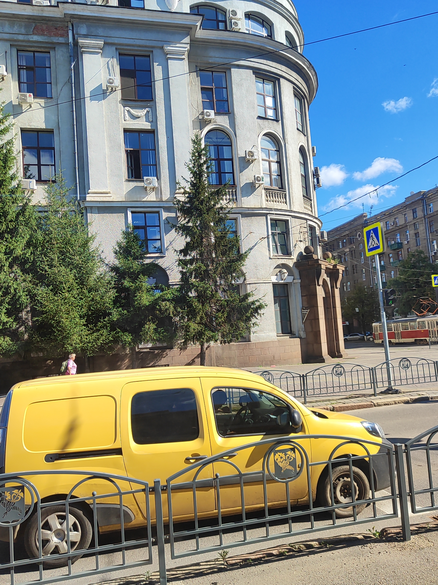A yellow panel van is parked in front of a large, light-colored building with arched windows and air conditioning units. The building has decorative pillars and is surrounded by evergreen trees. A person in a pink top and backpack is walking on the sidewalk. Across the street, a yellow pedestrian crossing sign and a traffic light are visible, along with a red and white tram. The sky is blue with scattered clouds. The scene appears to be an urban environment on a sunny day.