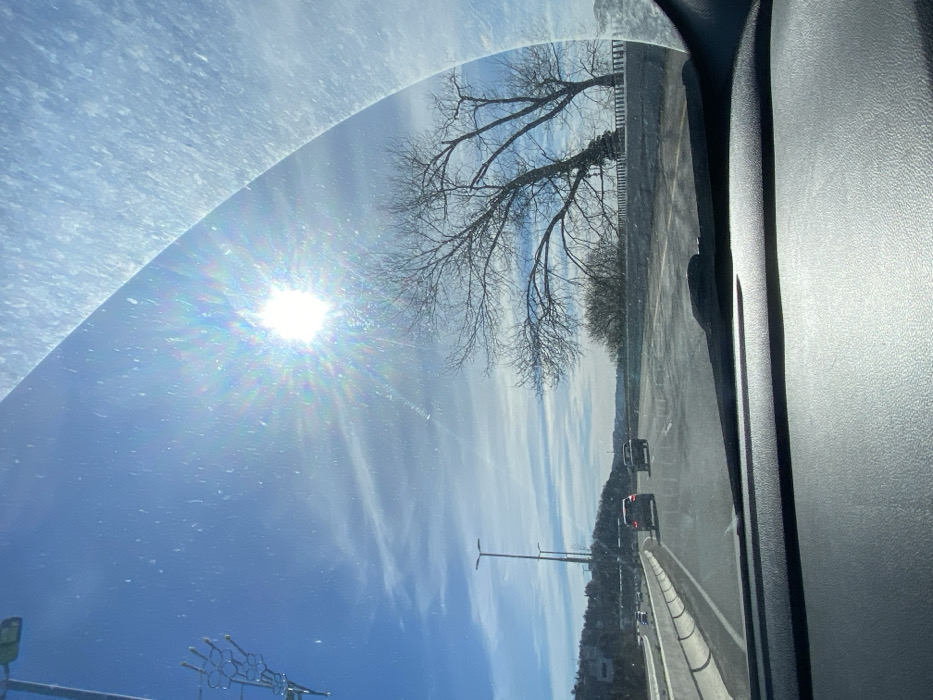 View from inside a car looking through the windshield, with bright sunlight refracting into a prism-like effect. A bare tree branch is visible against a blue sky streaked with light clouds. A highway stretches into the distance with moving cars. The road is lined with a concrete barrier on one side and a grassy hillside on the other. A few light poles stand along the road. The car's interior dashboard and door frame are partially visible in the foreground.
