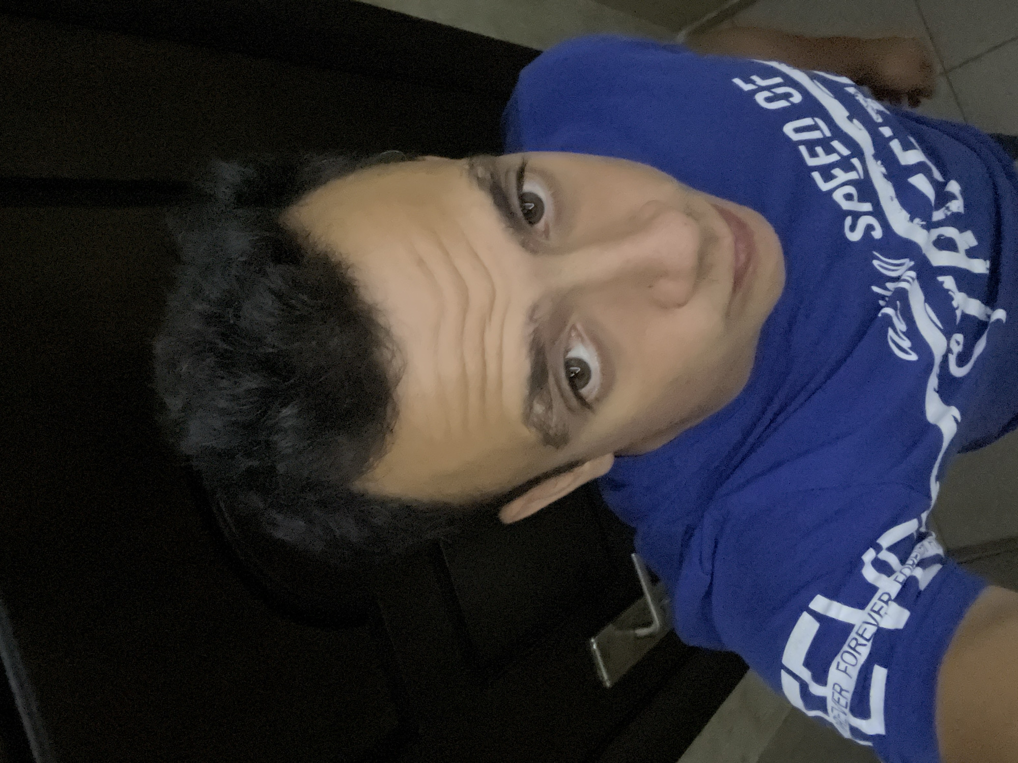 This is a vertical, close-up selfie of a person, likely male, who appears to be in their late teens or early twenties. They have dark, slightly wavy hair and light brown eyes. Their skin is smooth, and they are looking directly at the camera with a neutral expression.
The person is wearing a bright blue t-shirt with white text and graphics. The text "SPEED OF" is visible across the upper chest, with stylized white graphics accompanying it. Further down the sleeve, the words "FOREVER FOREVER" are repeated.
The setting appears to be indoors, with a dark background that could be a wall or a doorway. A sliver of a lighter surface, possibly a floor or furniture, is visible on the right side of the frame. The lighting suggests it could be daytime, but the indoor setting makes it difficult to determine the exact time or weather. There are no strong indicators of the specific location within Blagoevgrad, Bulgaria, beyond the subject's apparent presence there. The overall mood is casual and everyday.