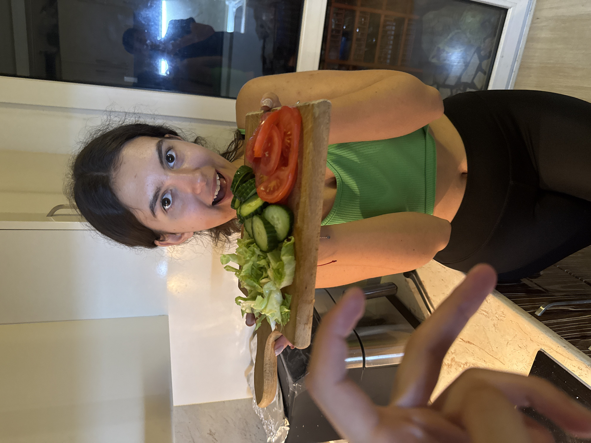 A young woman with dark hair is standing in a kitchen, holding a wooden cutting board with sliced tomatoes, cucumbers, and lettuce. She is wearing a green tank top and black leggings. Her mouth is open in a surprised or excited expression. In the foreground, a hand is partially visible, gesturing towards the cutting board. The kitchen appears to be modern, with light-colored cabinets and a granite countertop. A window in the background reflects a person. The overall scene suggests a casual moment in a home environment.