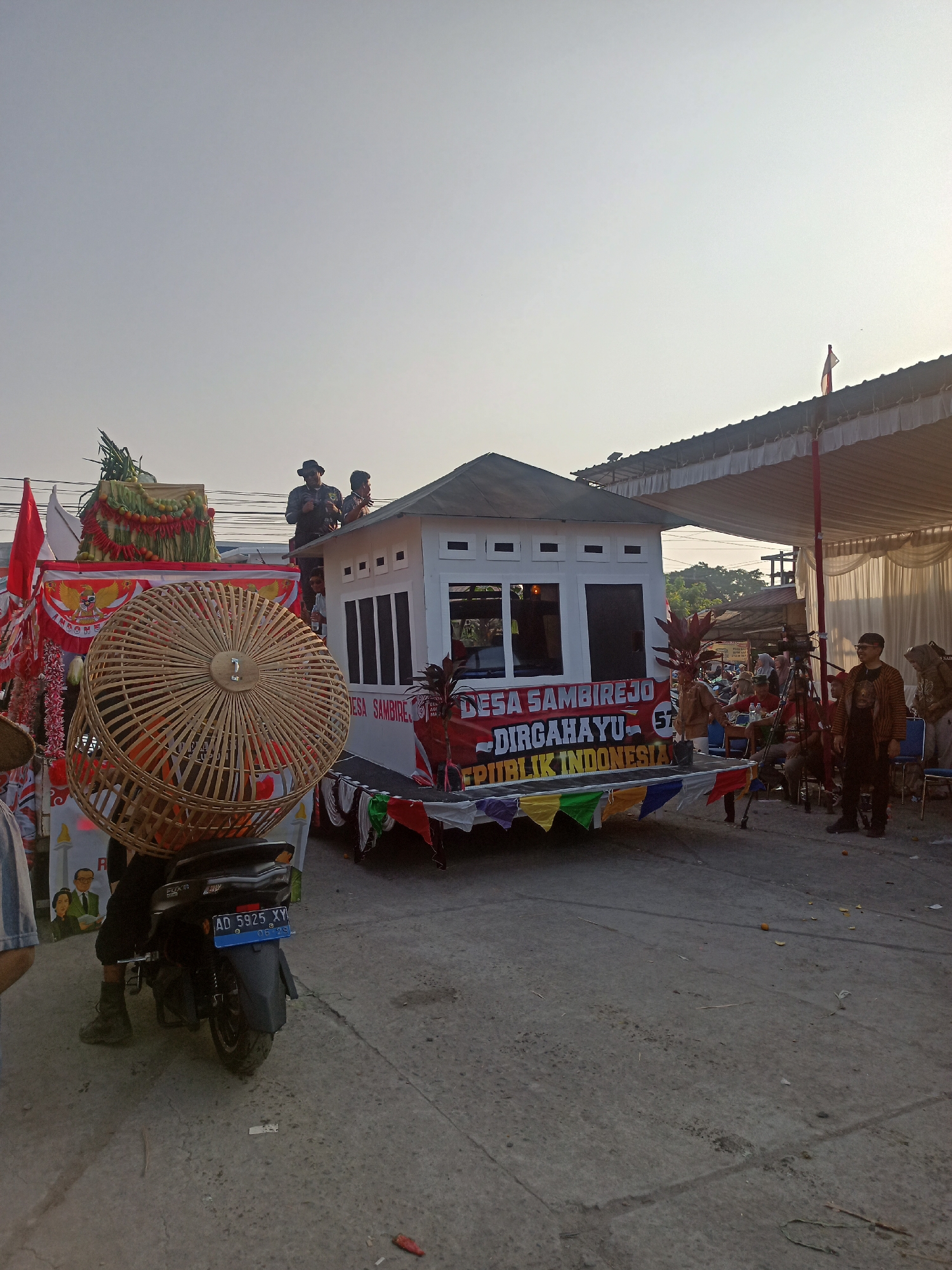 The image depicts a parade float in Ngrompongan, Indonesia, likely celebrating a national holiday. The float is a miniature house structure, adorned with banners that read "DESA SAMBIREJO DIRGAHAYU KE-57 REPUBLIK INDONESIA". Several people are visible on and around the float. Two individuals are standing on the roof of the miniature house, and others are positioned on a platform at the front. In the foreground, a motorcycle with a large, woven bamboo cage mounted on its rear is parked. The environment appears to be a street lined with tents and spectators in the background, suggesting an outdoor event. The sky is clear and bright, indicating it is daytime.