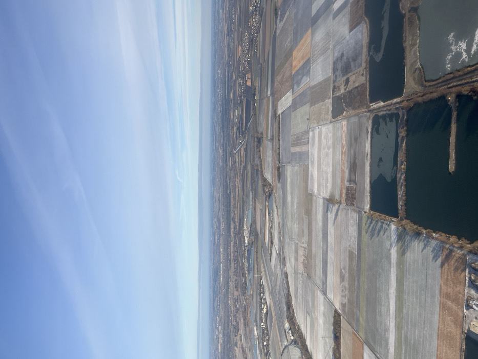 An aerial image captures a vast, flat, rural, and semi-urban landscape in the district of Erding, Germany, under a clear blue sky with minimal high-altitude cloud streaks. The scene is dominated by a mosaic of rectangular agricultural fields, many appearing light grey or white, consistent with frost, snow cover, or fallow earth, interspersed with patches of brown and darker tones. Numerous dark, irregularly shaped water bodies, characteristic of gravel pits, are prominent in the foreground and mid-ground, some exhibiting lighter, mottled patches indicative of partial ice formation.
A distinct, straight canal or large channel traverses the mid-ground horizontally, extending from the left to the right side of the frame and appearing to connect with other water features. Clusters of buildings, representing residential settlements or villages, are visible in the mid-ground and distant background, notably to the right and left. In the mid-left, an industrial complex comprising several buildings and associated water retention ponds is discernible adjacent to the main canal. Sparse, bare deciduous trees line some field boundaries and water edges, casting elongated shadows consistent with a low sun angle, suggesting a winter or late autumn season. No people or specific actions are discernible due to the high altitude perspective.