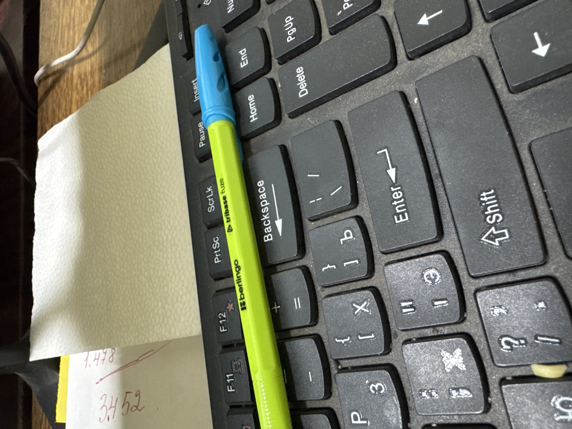 An overhead shot shows a bright green and blue pen resting on a black computer keyboard. The pen is positioned horizontally across several keys, including "Insert," "Pause," "ScrLk," "PrtSc," "F12," and "F11." The keyboard is dirty, with dust visible on the keys and around the edges. A white paper towel is partially visible in the upper left corner, with some red writing on it. The background is out of focus and appears to be a wooden surface with a white cable. The scene is well-lit, with a slight glare on the pen.