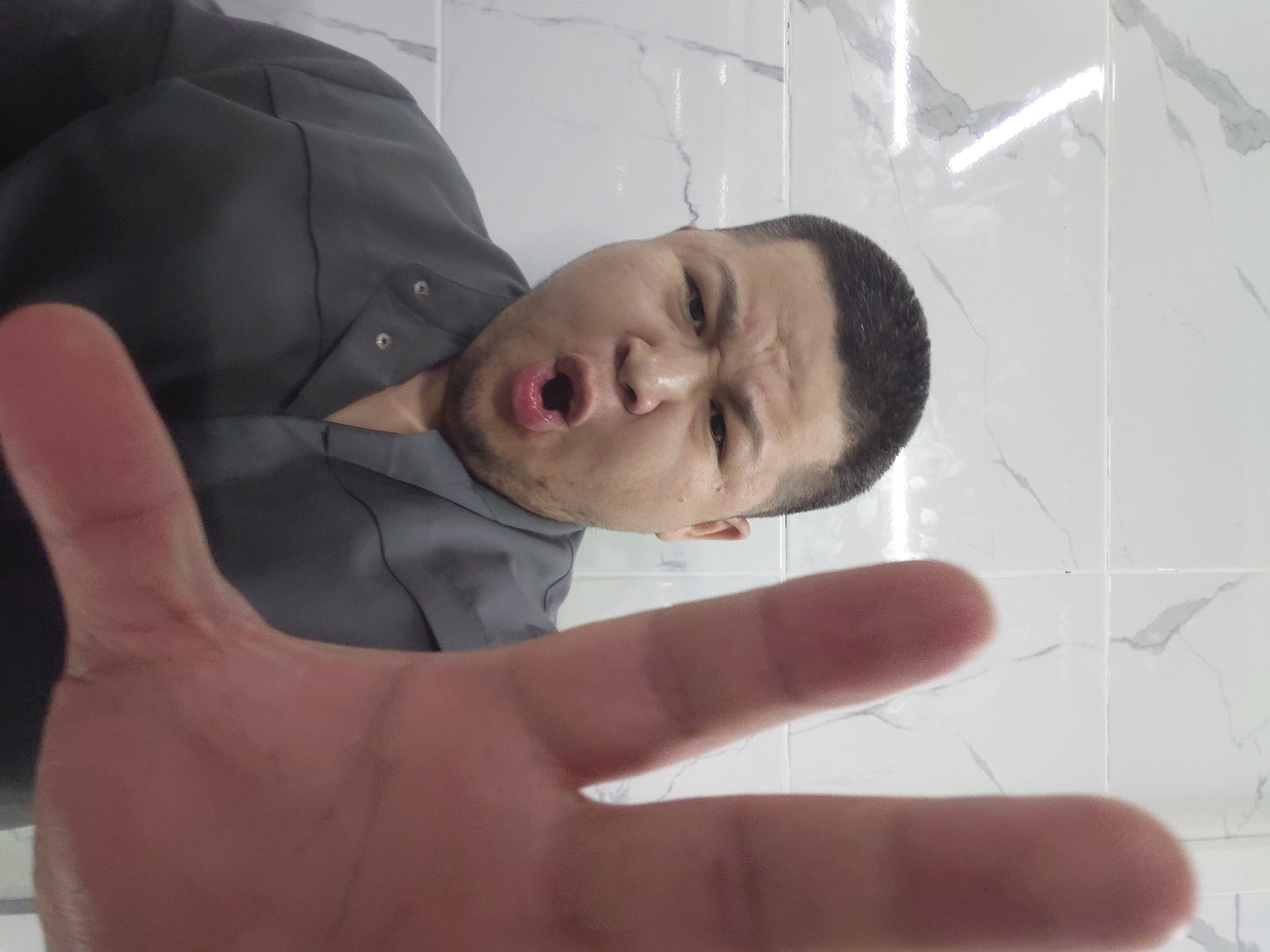 An extreme close-up selfie shows a man with short, dark hair and a surprised or angry expression. His mouth is open, and his eyebrows are furrowed. He is wearing a dark gray collared shirt. His right hand is held up to the camera, obscuring a large portion of the frame. The background consists of white tiled walls with gray veining, typical of a bathroom or kitchen. The lighting appears to be from overhead fluorescent lights. The setting is not identifiable as Aktau, Kazakhstan.
