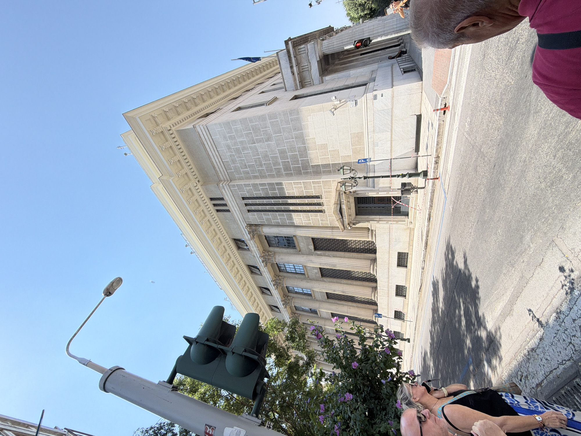 The image shows a view of the Bank of Greece building in Thessaloniki, Greece, from a street level perspective. A traffic light is visible in the foreground, attached to a pole. Next to the traffic light, there is a flowering tree with purple blossoms. Several people are standing on the sidewalk. The building itself is a large, neoclassical structure made of light-colored stone, featuring tall columns and intricate detailing along its roofline. The sky is clear and blue. The street in front of the building is paved with asphalt and marked with white lines. Shadows of trees are cast on the street. A flag is visible on the building's facade.