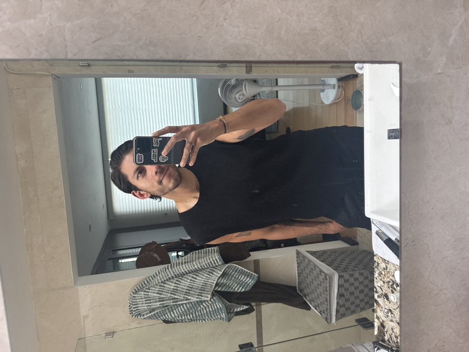 A man takes a selfie in a bathroom mirror. He wears a black t-shirt and a gold bracelet. The phone has stickers on it. Behind him is a window with blinds, a fan, a gray and white towel, and a woven storage cube. The bathroom has beige walls. A countertop and sink are visible along the right edge.
