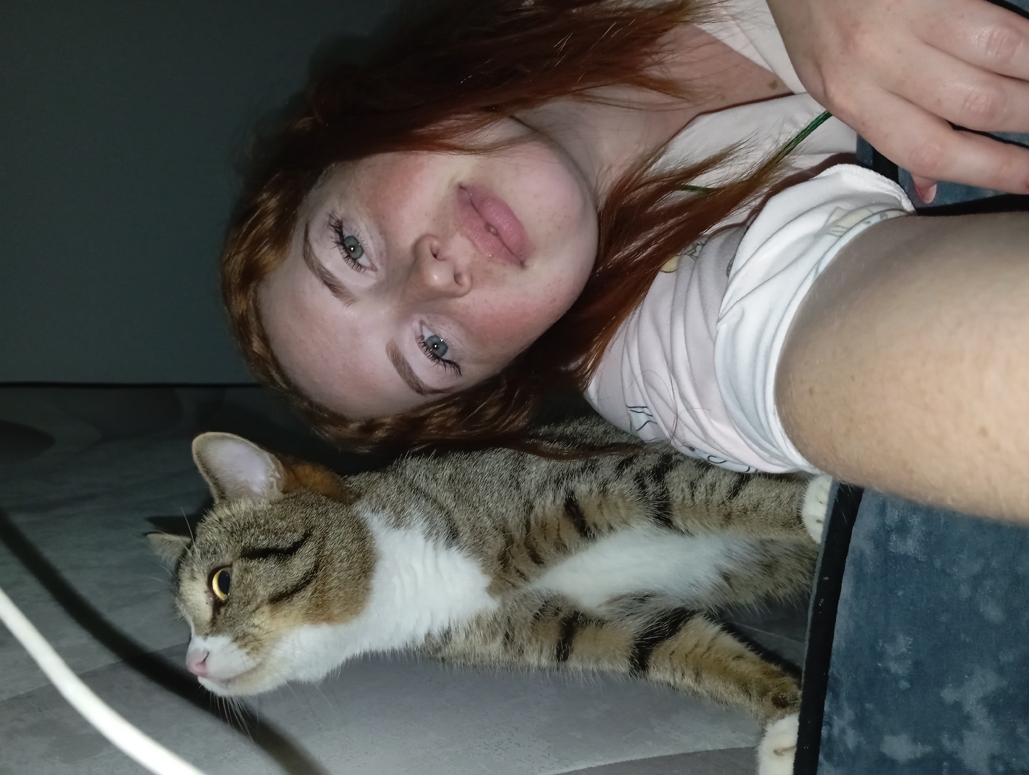 A young woman with auburn hair and freckled skin lies on a surface next to a tabby cat. The woman has light eyes and is wearing a white shirt with a light green strap visible. Part of her hand and a denim-covered surface are visible at the right. The cat is lying on its side with its paws extended. A white cable is visible in the lower left corner of the image. The lighting is subdued. The location, Yurgamysh, Russia, cannot be determined.