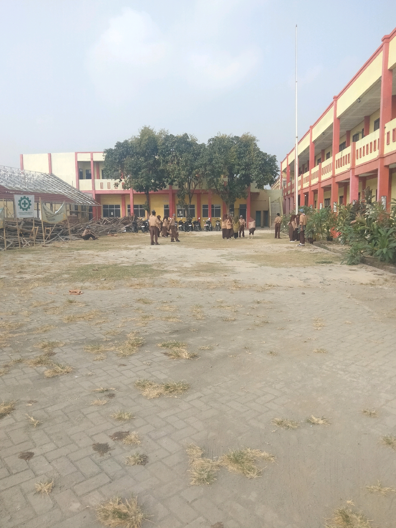 The image depicts an outdoor schoolyard scene in Suka Sari, Indonesia, with a multi-story building in the background. The building has a yellow facade with red trim and multiple windows and balconies. In the foreground, the ground is paved with interlocking bricks, with patches of dry grass and dirt. Several individuals, appearing to be students in uniform, are present in the middle ground, some standing in groups and others walking. To the left, there is a partially constructed or dismantled structure made of bamboo and corrugated metal, with a sign bearing a green safety emblem. A tall flagpole stands near the center of the image. The sky is overcast. Motorcycles are parked to the left of the building, near an entrance. The overall impression is a somewhat unkempt school grounds with ongoing construction or demolition activities.