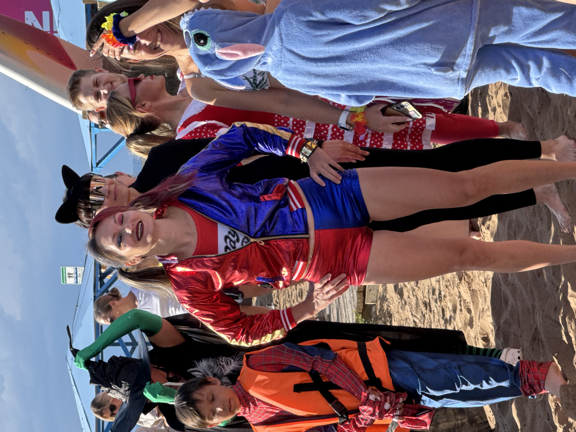 A group of people are posing for a photo on a sandy beach. The image is taken from a high angle, looking down at the subjects. The people are dressed in costumes, including a person in a blue Stitch costume, someone dressed as Harley Quinn, and a child in a Spiderman costume with a life vest.
The scene appears to be a lively event, possibly a costume party or a themed gathering. The people are smiling and appear to be in good spirits. The time of day is likely afternoon, as indicated by the strong shadows and bright sunlight. The weather is clear and sunny.
Notable details include the variety of costumes, suggesting a fun and playful atmosphere. The sandy beach setting implies a recreational activity or event taking place near the water. The people are positioned closely together, some of them sitting or lying down.
There is a sign visible on a blue structure in the background with text that is difficult to read clearly, but it appears to have some pictogram.
The overall impression is of a joyful and energetic gathering on a beautiful day.