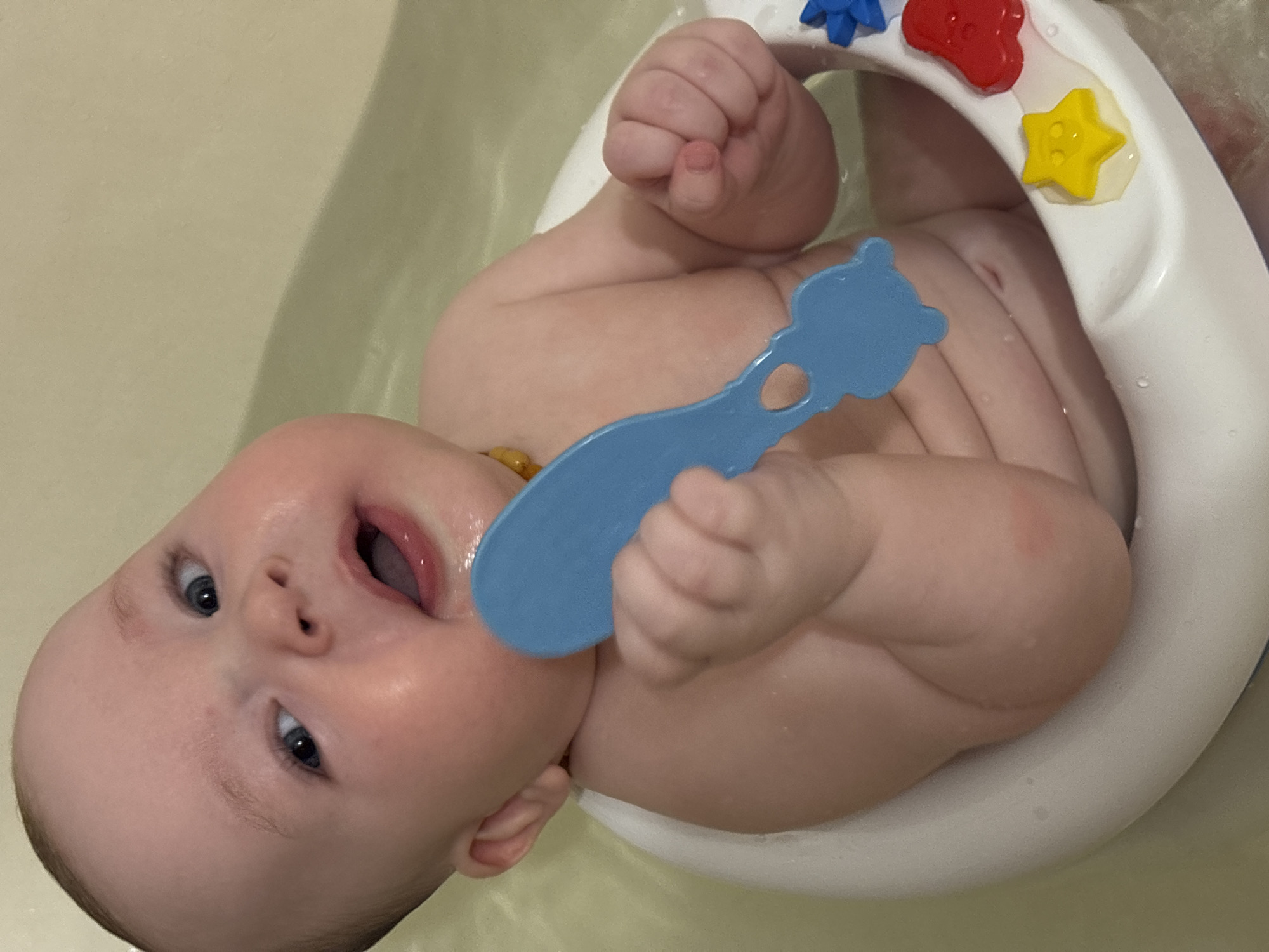A baby lies in a white plastic infant tub, partially submerged in water. The baby has fair skin, blue eyes, and a partially open mouth. The baby is holding a blue, bear-shaped teething toy to its mouth. The tub has a curved rim with colorful plastic shapes attached. The surrounding environment suggests a bathroom setting. The image does not provide sufficient information to determine a location in the city of Марковский, Russia.