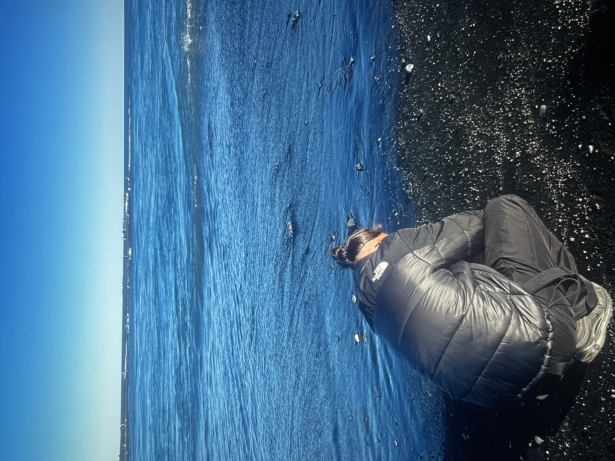 A woman wearing a black puffer jacket and dark pants is sitting hunched over on a black sand beach. The scene includes a blue sky on the left and an ocean with waves. Small white objects, likely pebbles, are scattered across the black sand. The location is a black sand beach in an unknown city.
