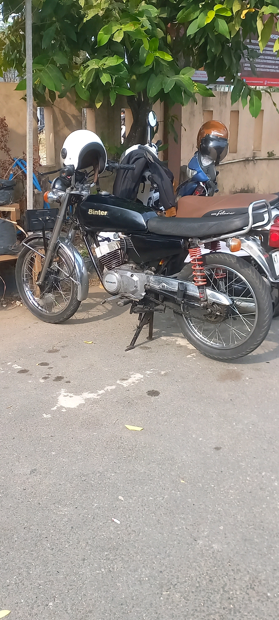 The image shows a street scene in Samarinda, Indonesia, with two motorcycles parked side-by-side. The foreground features a black motorcycle with a "Binter" logo on the fuel tank and "Kawasaki" branding on the engine. It has a brown seat with white lettering that reads "flame". A white helmet with a black stripe is resting on the handlebars. Behind this motorcycle is a blue scooter with a brown helmet resting on its handlebars. The motorcycles are parked on an asphalt road with some debris and oil stains on the surface. In the background, there is a wall and lush green foliage from trees. The lighting suggests it is daytime, likely outdoors in a built-up area.