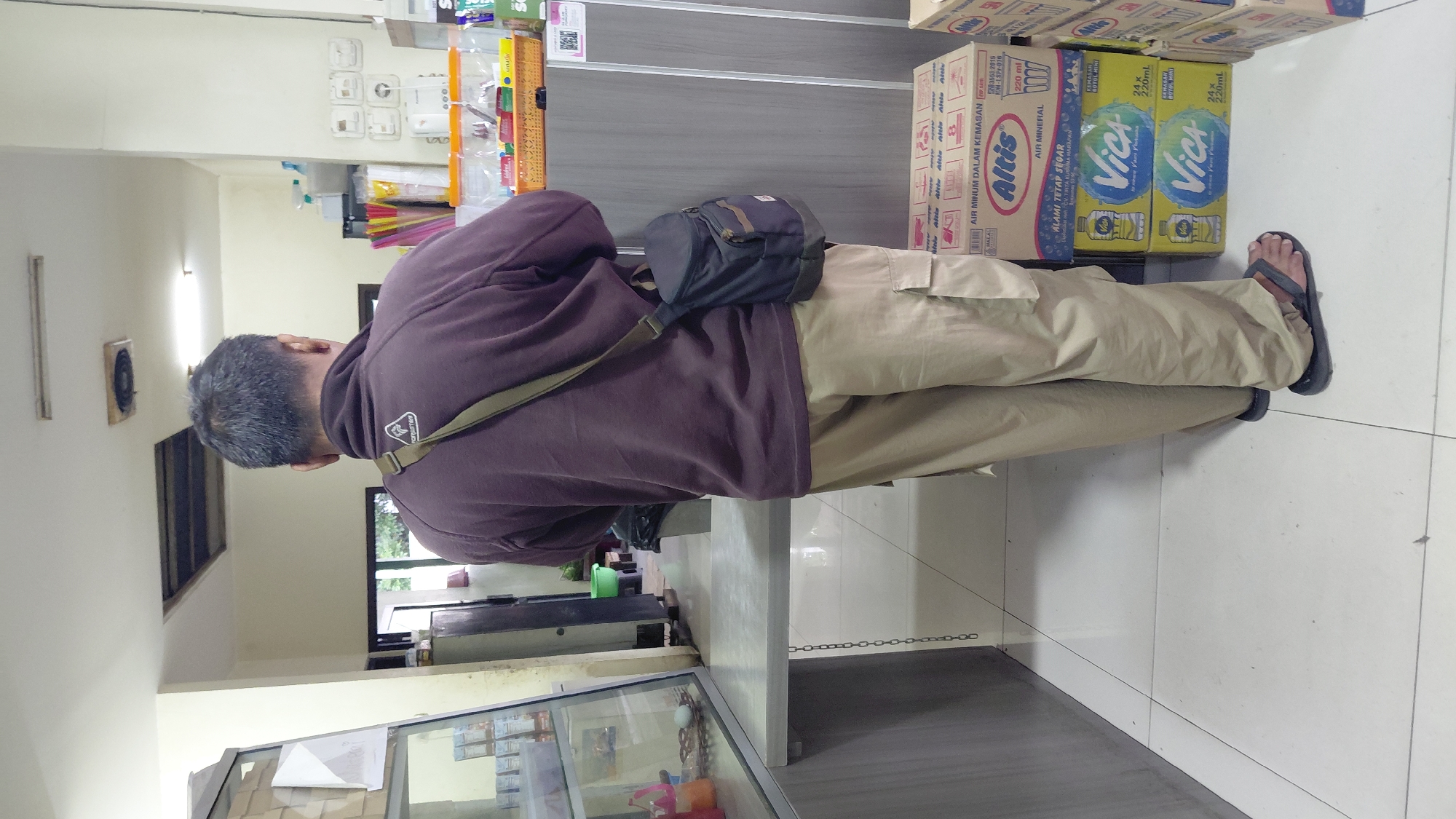 A medium shot captures a man from the back, standing in a dimly lit room that appears to be a small shop or convenience store in Rejasari, Indonesia. The man, wearing a dark purple long-sleeved shirt and light brown cargo pants, has a dark bag slung across his right shoulder. He is facing away from the camera, toward a counter with various items displayed on shelves behind it. In the foreground, to the right of the man, are several stacked boxes of bottled water, some with labels indicating "Altis" and "Vica." The floor is tiled with large white squares. The overall environment suggests a retail setting with goods for sale.