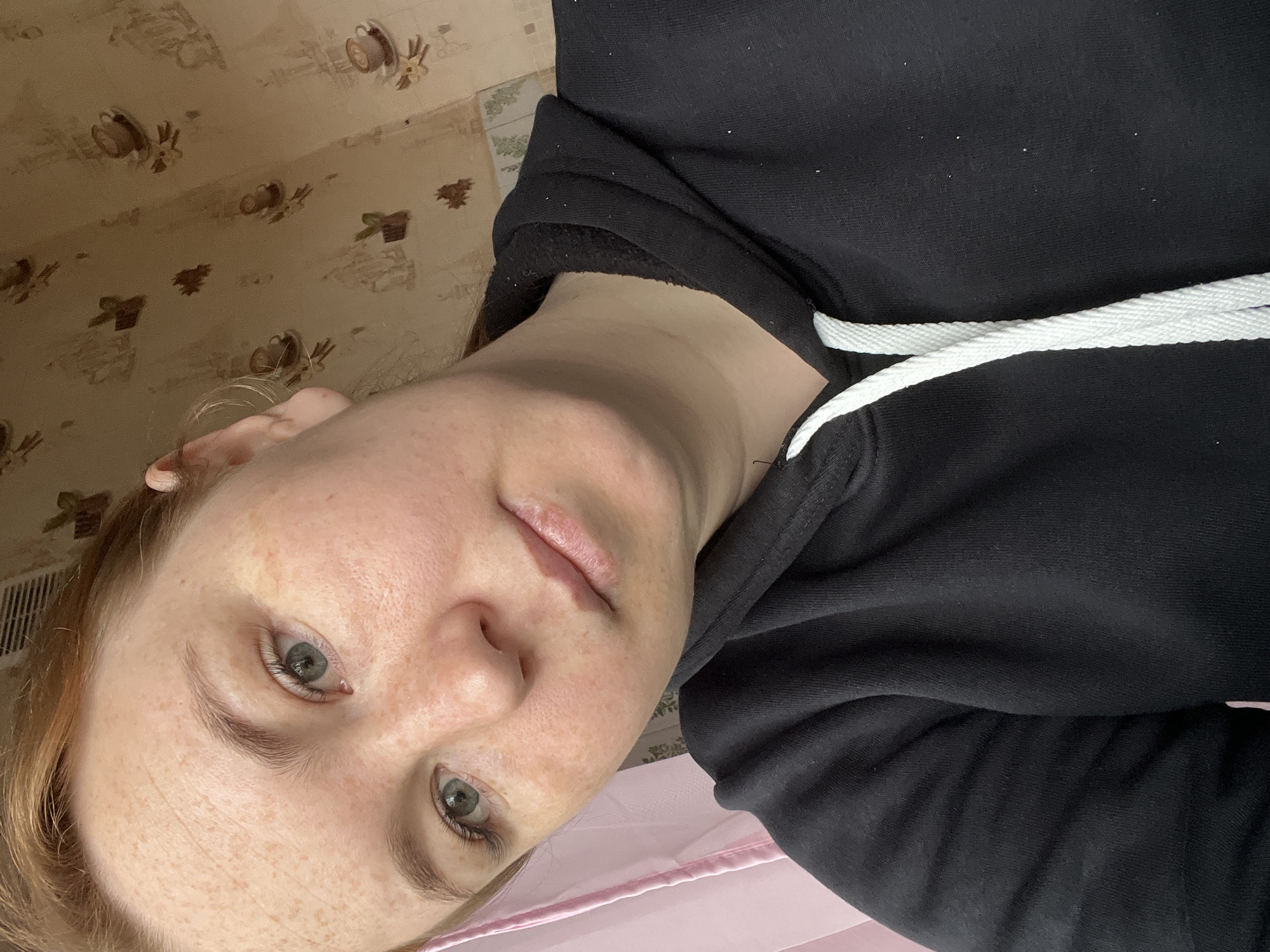 The image is a close-up, rotated selfie of a person, likely a woman, taken indoors. She has light reddish-brown hair, fair skin with noticeable freckles, and light-colored eyes, appearing greenish-grey, looking directly at the camera. Her expression is neutral, with lips slightly parted. She wears a black hoodie, visible across her upper body, featuring a prominent white drawstring. Small white specks, possibly dust or lint, are visible on the black fabric.
The background primarily features a light brown or beige wallpaper with a repeating pattern of brown coffee cups and abstract shapes, possibly leaves. Parts of the wallpaper show signs of wear or peeling. A sliver of light pink fabric or an object is visible in the lower right portion of the frame. The lighting is soft and even, typical of indoor settings. The scene depicts a casual indoor environment, possibly a home in Chaikovsky, Russia. No visible text is present. The activity is a self-portrait.