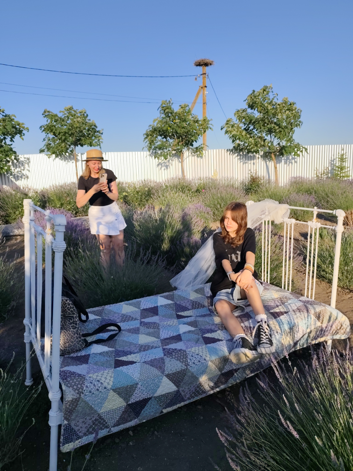 In Nechaiane, Ukraine, a woman wearing a straw hat and a white skirt stands in a lavender field, holding a smartphone and taking a photo. She is positioned behind a white metal bed frame, which is covered with a colorful patchwork quilt. To the right of the frame, a young girl with brown hair sits on the bed, wearing a black t-shirt and denim shorts. She looks towards the side with a thoughtful expression.
Behind the subjects, a white picket fence stretches across the background, with several small trees planted in front of it. In the distance, a stork's nest sits atop a utility pole. The scene is bathed in the warm, golden light of late afternoon or early evening, suggesting a pleasant, clear day. The presence of the lavender and the clear sky indicate an outdoor setting, likely a garden or a rural landscape. The overall mood appears relaxed and idyllic, with the woman engrossed in capturing the moment and the girl enjoying the tranquil surroundings. The image captures a peaceful, summery scene with a touch of whimsy provided by the bed set up in the lavender field.