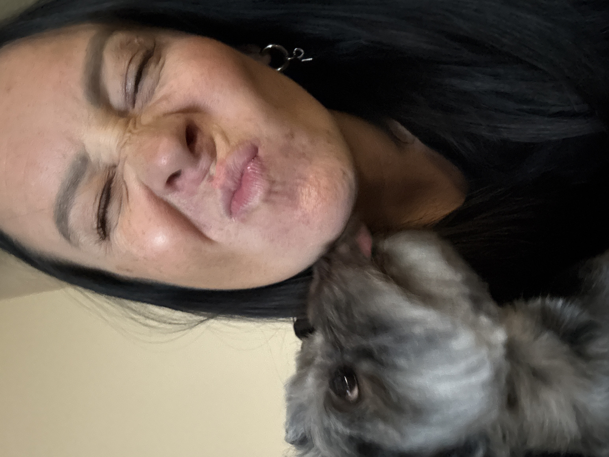 The media file is a close-up, horizontally oriented photo of a person and a dog. The person is in the foreground, with their eyes closed and their face contorted in a playful expression, perhaps a grimace or a laugh. Their mouth is slightly open, and their tongue appears to be visible. They have dark hair and are wearing an earring. The dog, a small, gray, fluffy breed, is in the process of licking the person's face, specifically near their mouth. The dog's eye is visible and appears to be looking towards the camera. The background is a plain, light-colored wall, suggesting an indoor setting. The lighting is soft and appears to be artificial, casting a warm glow on the subjects. The overall mood of the image is affectionate and lighthearted, capturing a moment of playful interaction between a human and their pet. There is no visible text in the image.