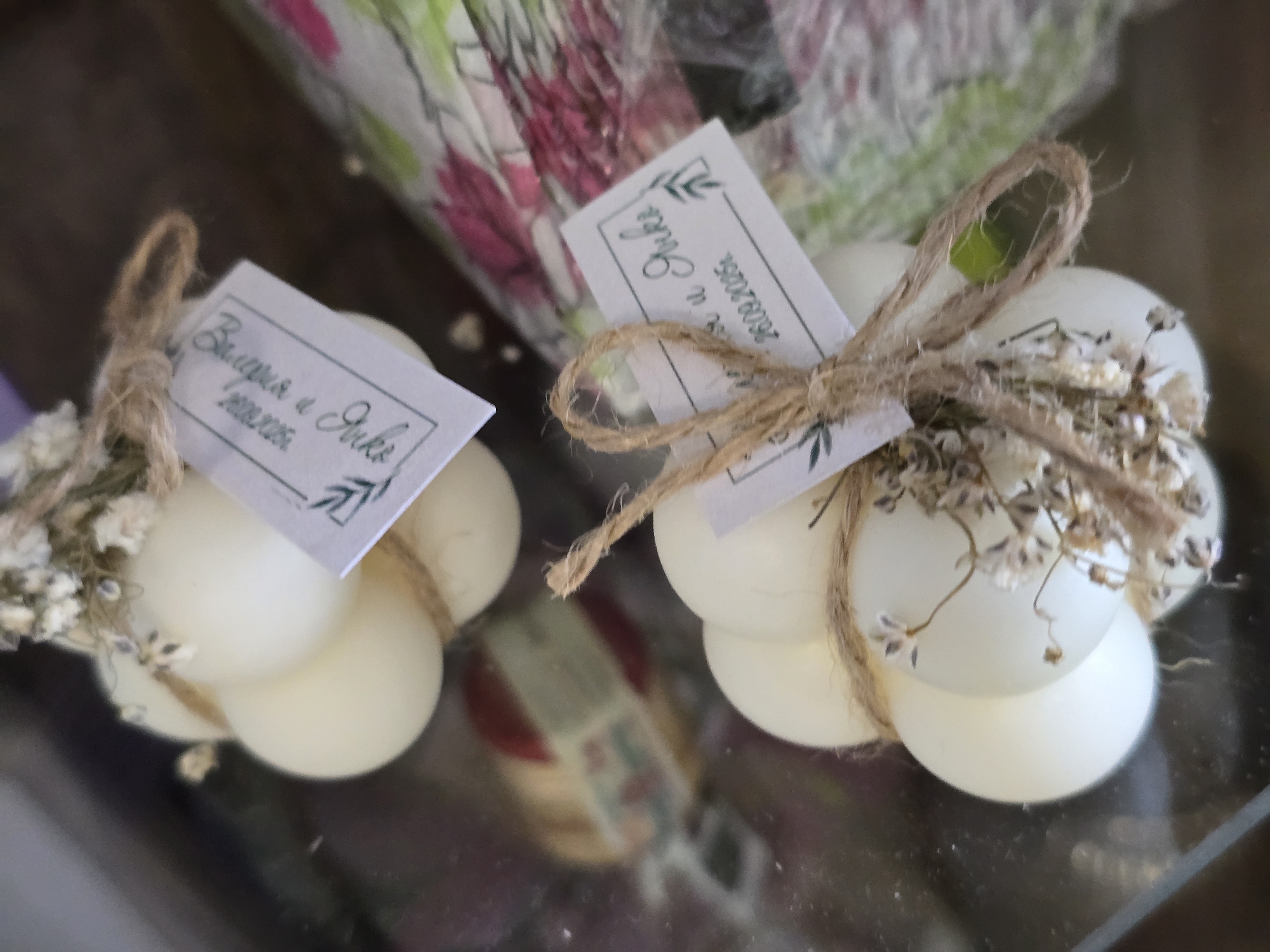 The image displays two decorative white objects, likely candles, each formed from a cluster of spherical shapes. Both objects are adorned with natural jute twine, tied into bows, and embellished with small sprigs of dried white flowers, possibly baby's breath. Each item also features a small rectangular white tag, personalized with text and a green leafy design.
On the left tag, the visible text in a calligraphic script reads "Валерия и Юли" (Valeriya and Yuli), followed by the date "19.09.2006". The tag on the right also displays "Валерия и Юли" (Valeriya and Yuli) with the date "28.09.2006" below it, though the text is oriented vertically and partially obscured by the twine.
The objects rest on a reflective, dark surface, likely glass, which shows faint reflections of the items and the background. The background is softly blurred but appears to be a patterned material, possibly fabric or paper, exhibiting shades of pink, green, and white. The setting suggests an indoor environment, possibly a display for gifts or favors at a celebratory event, such as a wedding or anniversary, indicated by the personalized names and dates. The overall impression is one of thoughtfulness and celebration.
