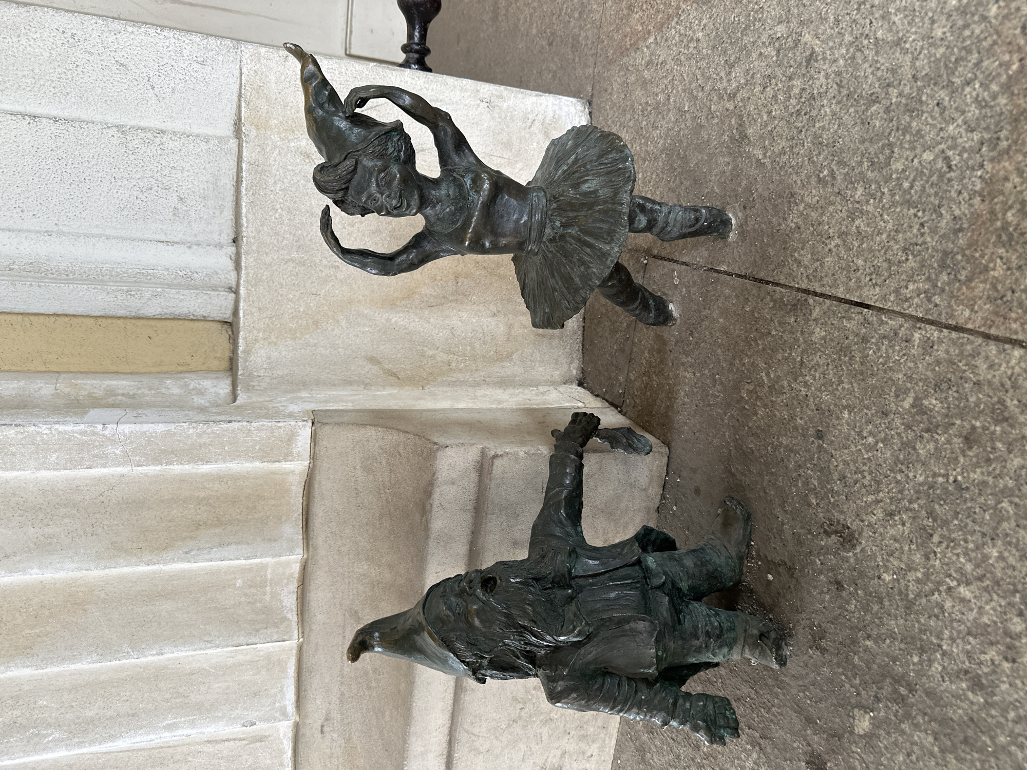 The image depicts two small bronze statues of gnomes, known as Krasnale, in Wrocław, Poland. One gnome is a ballerina in a tutu, posed in a dancing position on a stone staircase. The other gnome wears a pointed hat and is lying prone on a paved surface. The statues are tarnished and appear weathered. The environment consists of the stone staircase and a paved ground.