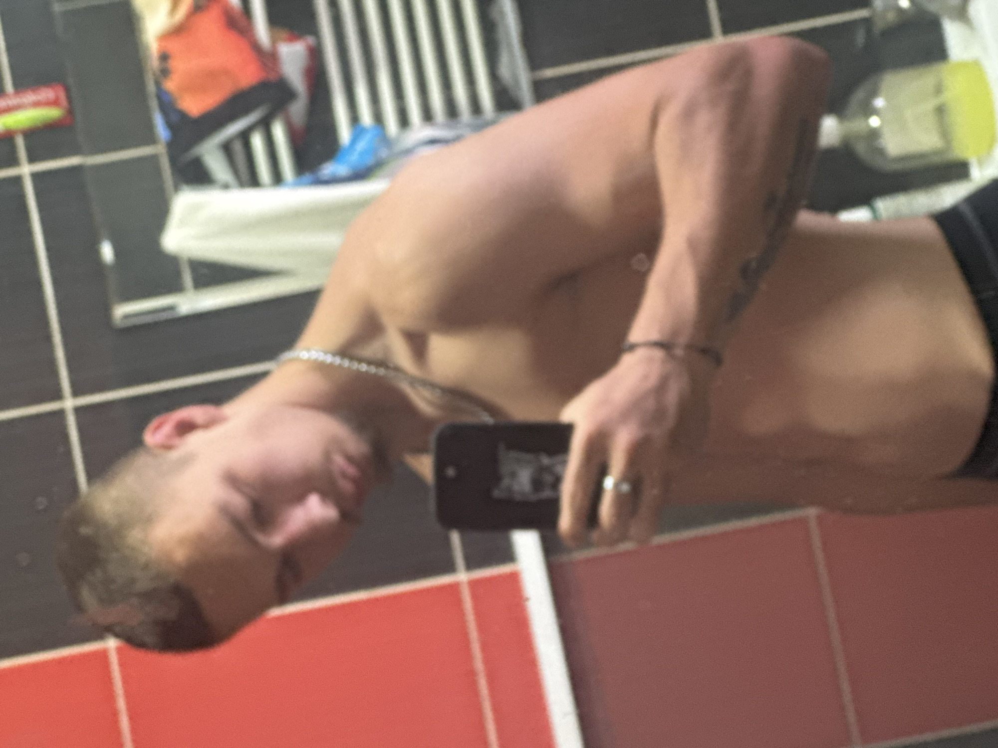 An overhead-oriented mirror selfie captures a shirtless young adult male in a bathroom in Vetren Dol, Bulgaria. The man, with short dark hair, is seen from the chest up, facing slightly away from the camera's perspective, looking towards his reflection. He holds a black smartphone in his right hand to take the photo, with a silver ring visible on his right ring finger. A silver chain is worn around his neck, and tattoos are visible on his left forearm.
The bathroom features dark grey/black tiled walls with white grout lines in the upper section and bright red tiles with white grout lines in the lower section, separated by a white border. In the mirror's reflection, behind the man, a white towel hangs over a rack. Various bathroom items are visible, including a bottle with a yellow cap and a small red and green object, possibly a product tube, on a shelf. No discernible text is visible on any of the products or items in the reflection due to blurriness. The time of day or specific weather conditions are not evident from the indoor setting.