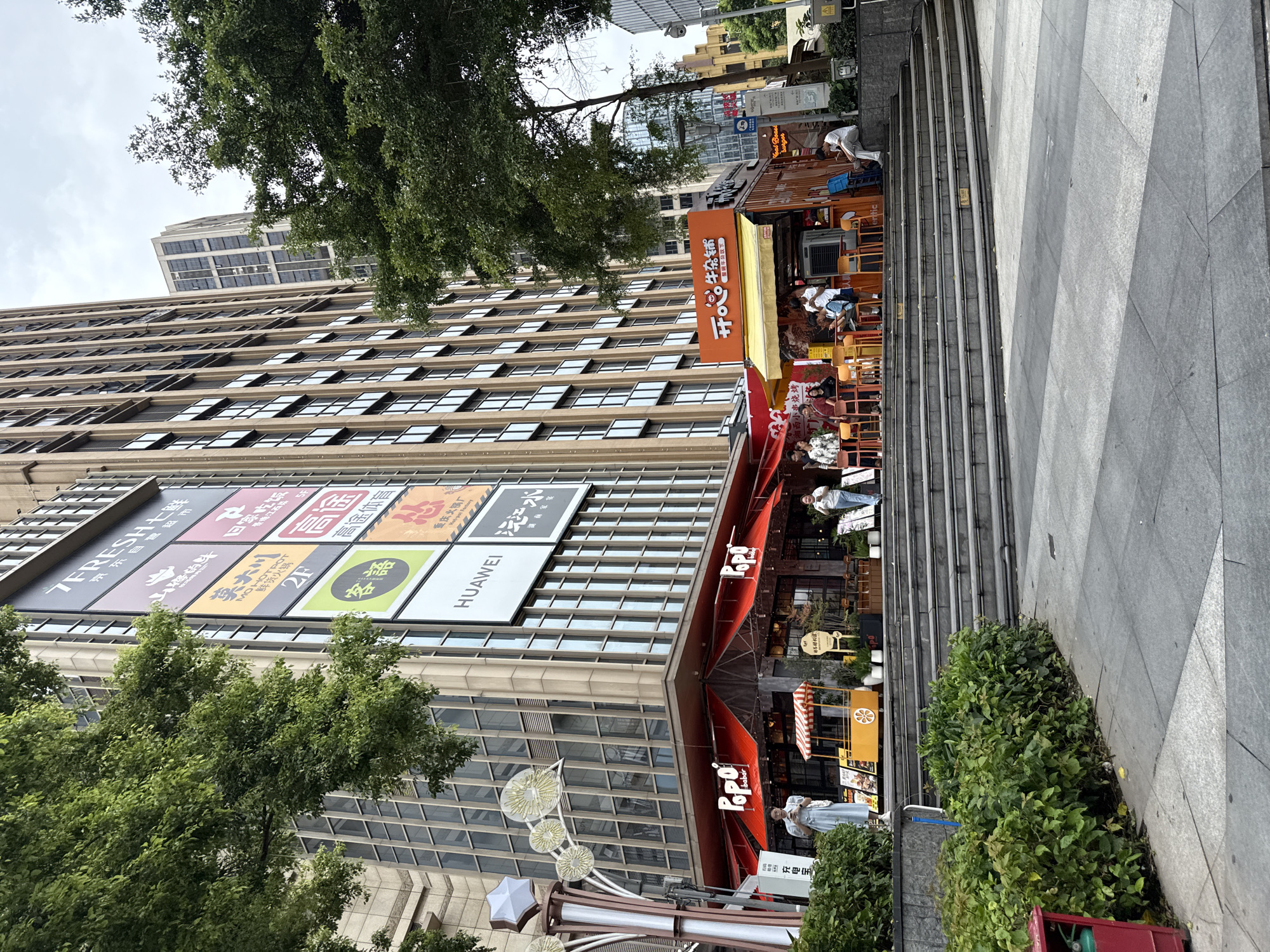 The image captures a bustling street scene in Haizhu District, China. A tall, modern building dominates the background, its facade covered in large advertisements and windows. In the foreground, a wide set of stone stairs leads up to a commercial area featuring shops and food stalls. Several people are visible, some sitting at outdoor tables, others walking or standing. Lush green trees frame the scene, adding a touch of nature to the urban environment. The overall impression is one of a vibrant and active urban hub.