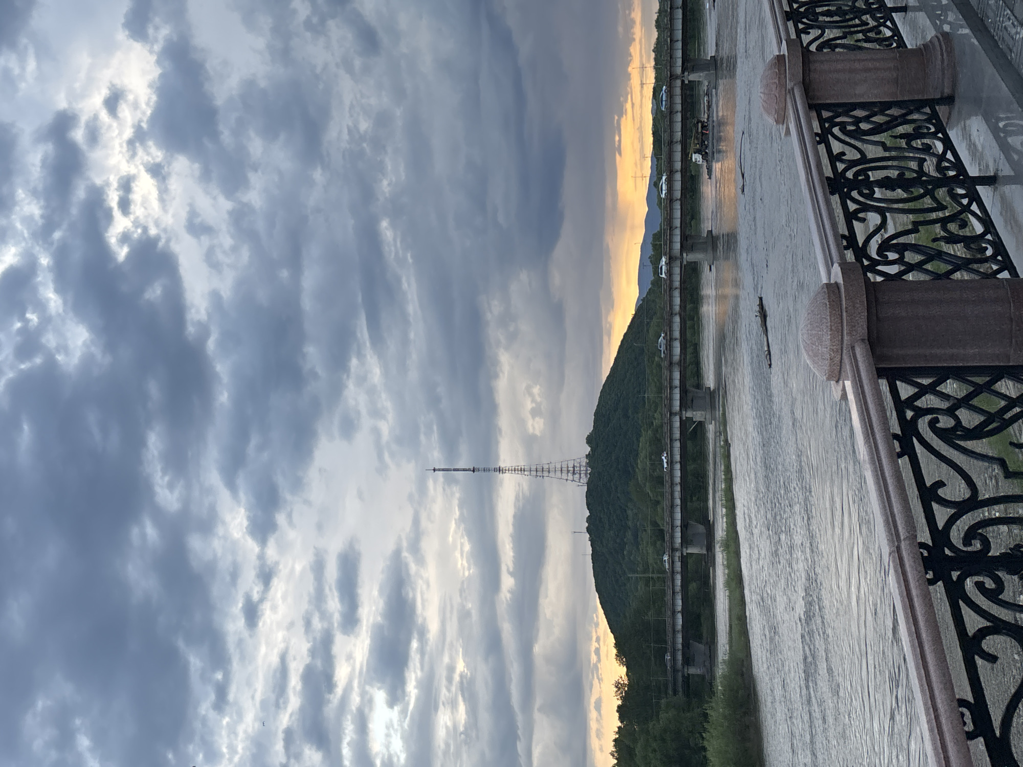A wide shot captures a cloudy sky over a river, a hill with a communications tower, and two bridges. In the foreground, a decorative metal railing with granite posts overlooks the choppy, gray river. Beyond the river, a long bridge with multiple cars is visible, followed by another bridge with a truck. The hill, covered in green trees, rises in the background, topped by a tall, skeletal communications tower. The sky is filled with dramatic, dark gray clouds with patches of lighter gray and yellow peeking through at the horizon, suggesting either sunrise or sunset.