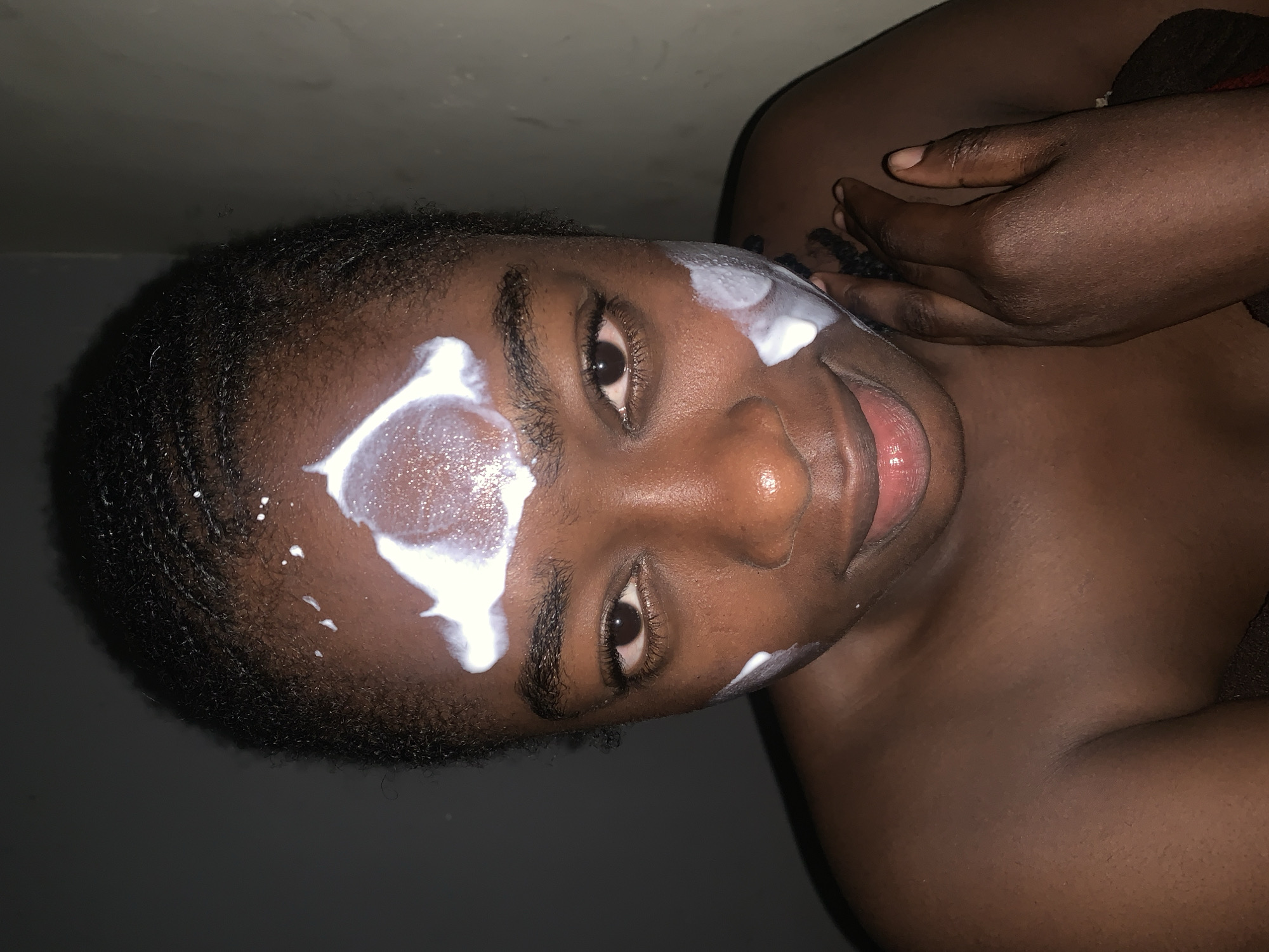 A young, dark-skinned individual, likely female, is depicted in a close-up portrait from the chest up, positioned indoors in the city of Rafin Par, Nigeria. She has short, dark, tightly textured hair, possibly in small braids or cornrows. Her brown eyes are directed forward, engaging the viewer, and her lips are slightly parted in a gentle smile. White cream or product is visibly applied to her face: a significant, somewhat crescent-shaped dollop is present on her forehead, with smaller, scattered dots above it, and additional patches are located on her right and left cheeks. Her right hand is visible on the lower right side of the frame, resting near her chin or neck area. The subject appears bare-shouldered. The background is dark and largely indistinct, with subtle hints of a textured wall or surface on the right, indicating an intimate, low-light indoor environment.