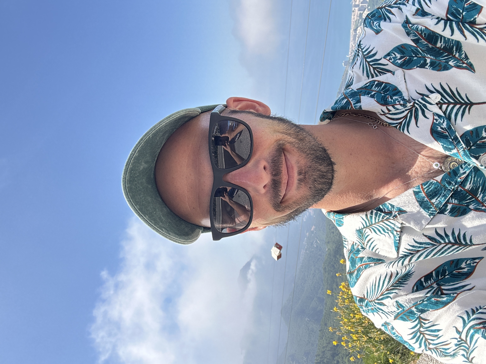 A man is captured in a selfie from the chest up, wearing a green baseball cap, dark sunglasses, and a white short-sleeved shirt with a tropical leaf pattern. His beard is trimmed, and he has a slight smile. The background reveals a landscape with mountains, a body of water, and a distant city. The sky is blue with scattered clouds. In the reflection of his sunglasses, a second image of the man holding the camera is visible, along with a street. The location is identified as Brajici, Montenegro.