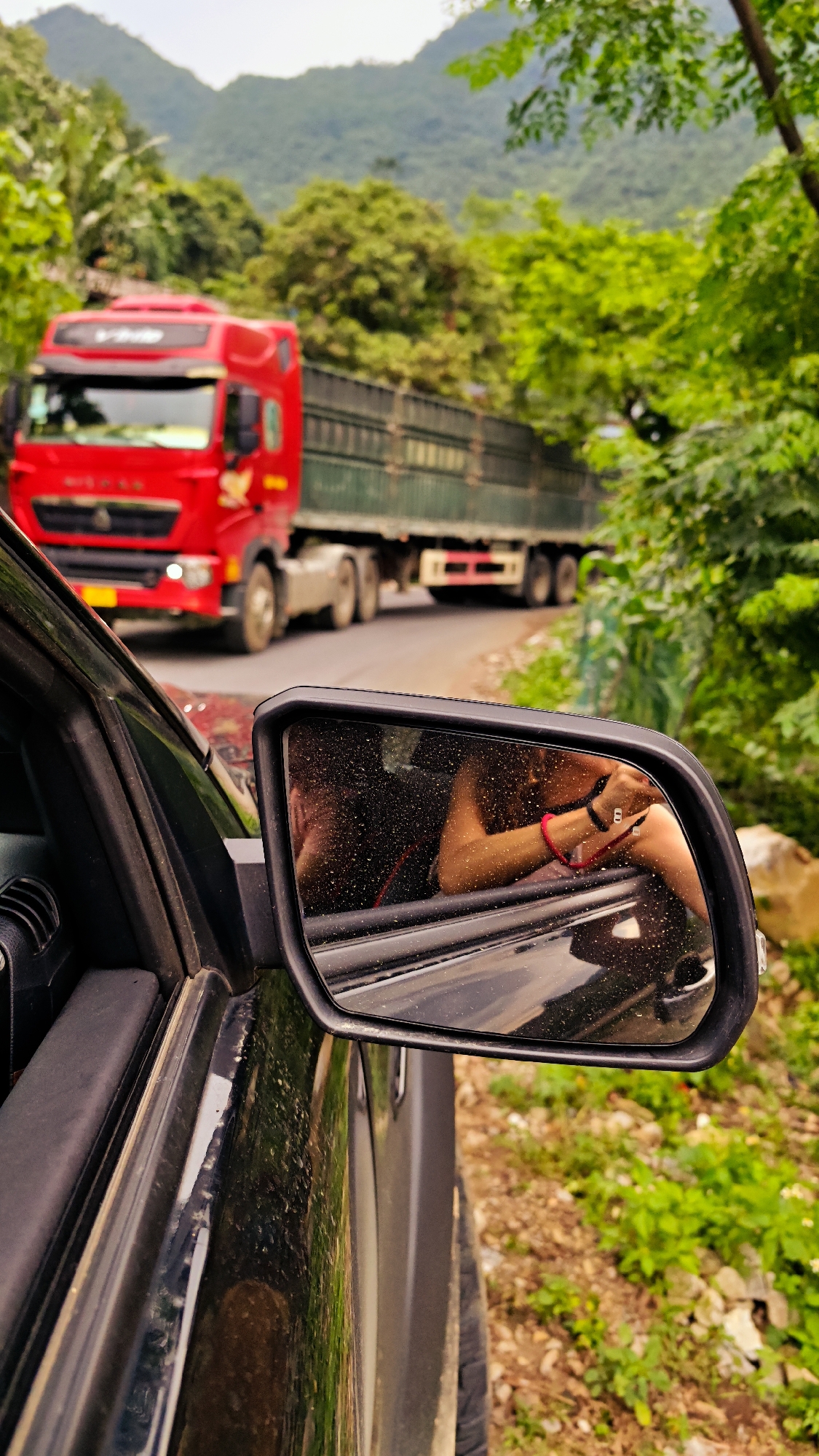 The image, taken from the perspective of a driver in a vehicle at Cán Tỷ Commune, Vietnam, shows a red semi-trailer truck traveling in the opposite direction on a narrow road lined with lush vegetation and mountains in the background. The side mirror of the vehicle reflects the arm of a passenger. The scene suggests a rural or countryside location within Cán Tỷ Commune.