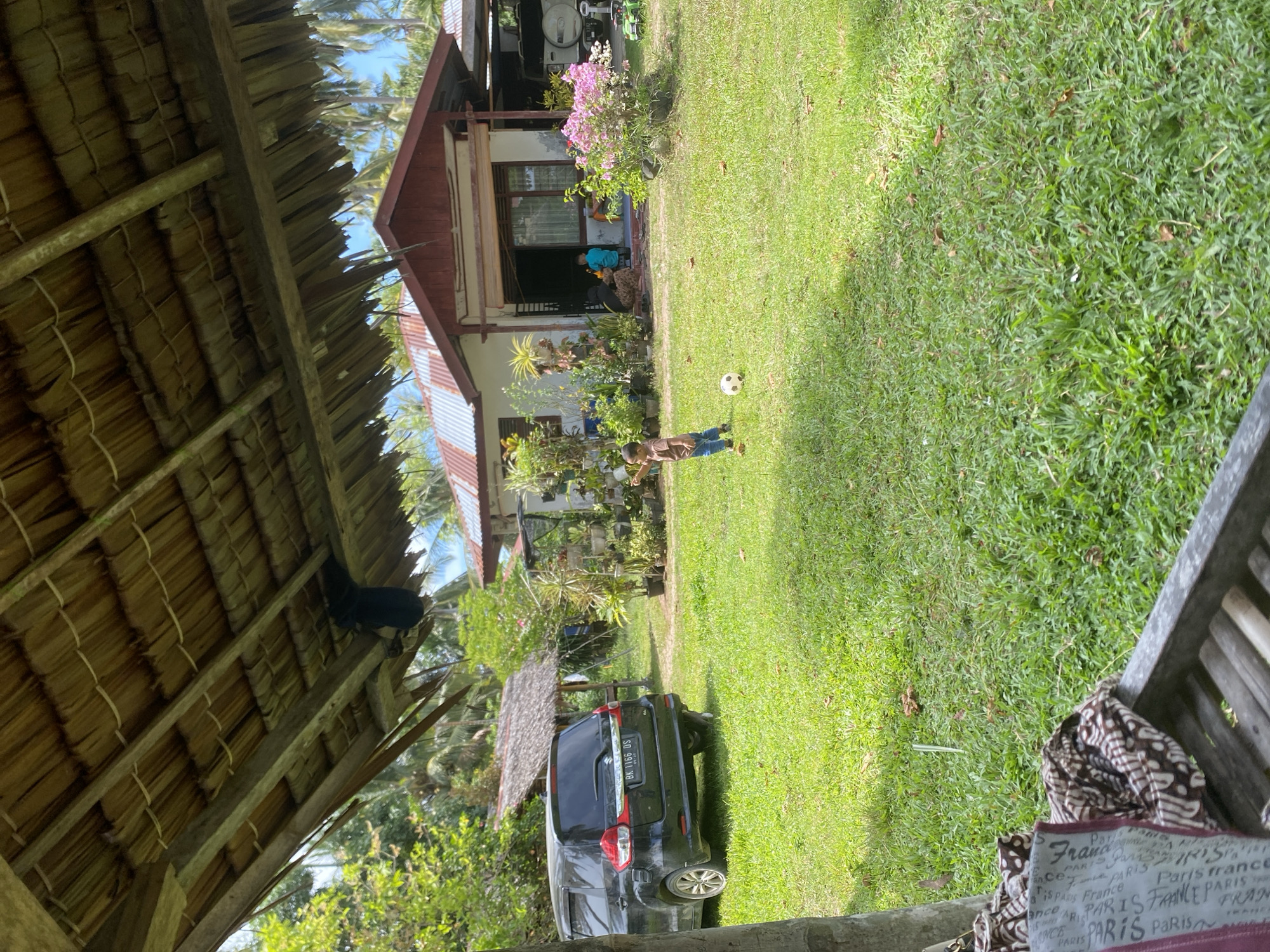 The image appears to have been taken in a rural or suburban setting, potentially in Payadua, Indonesia, as evidenced by the license plate visible on a dark-colored car, and the presence of tropical vegetation. The foreground features the interior of a thatched-roof structure. Outside, a young person is playing with a soccer ball on a green lawn in front of a one-story house with a reddish-brown roof. A couple of individuals are seated on the porch of the house. There are trees and additional plants around the yard, and the sky is partially visible beyond the house.