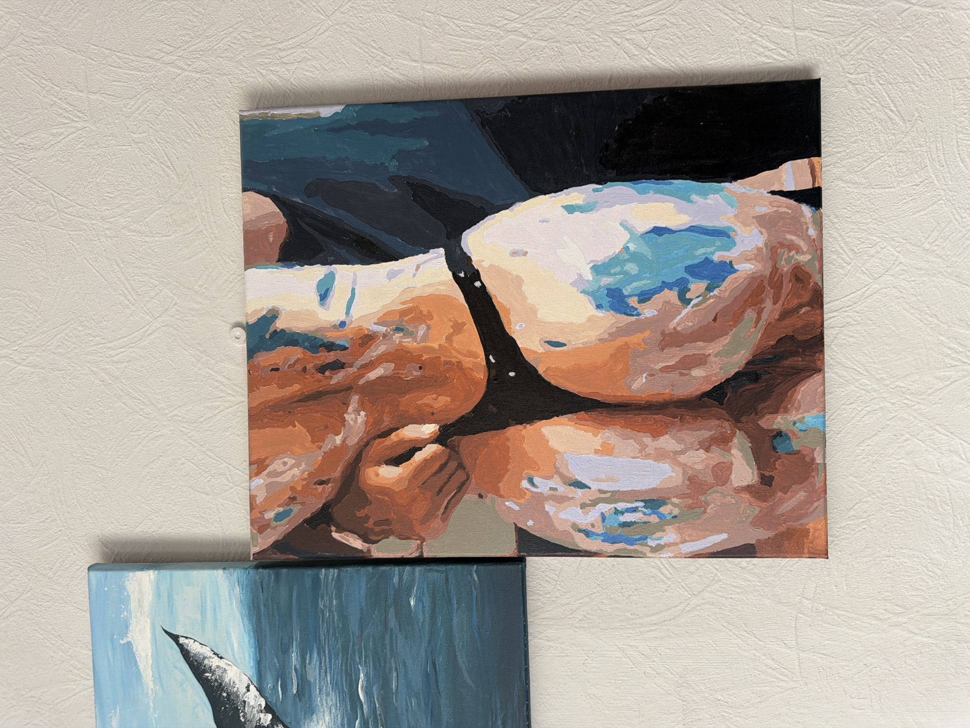 The image shows two square paintings hanging on a textured, off-white wall. The upper painting depicts a body of water with figures swimming, rendered in a blocky, impressionistic style with blue, brown, and white paint. The lower painting, partially visible, shows a blue and white image, potentially a whale or wave. The setting is presumably an interior space in an unknown location, with no evidence to place it specifically in Moscow, Russia.