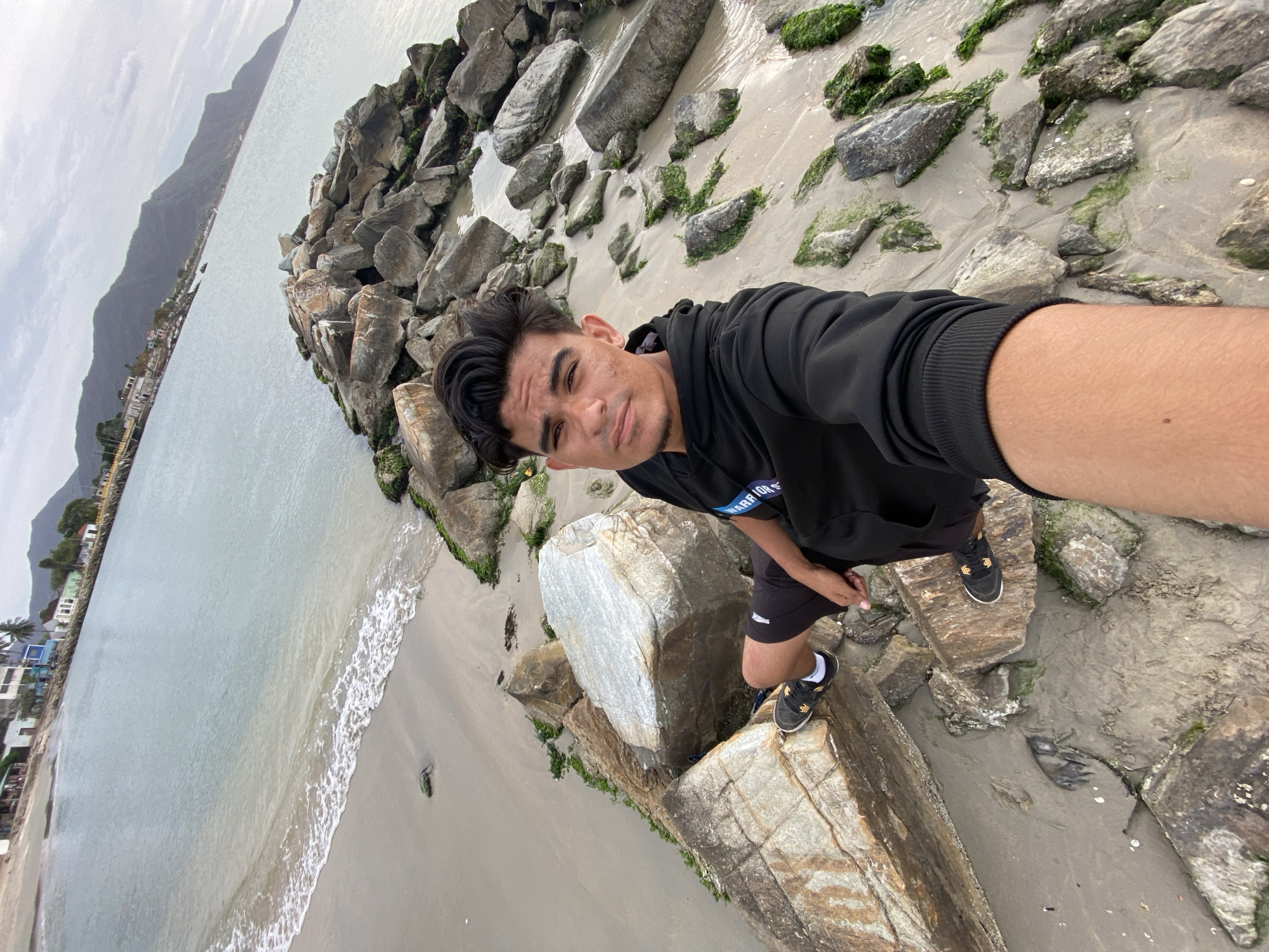 The scene depicts a young adult male taking a selfie while situated on a rocky breakwater in Juan Griego, Venezuela. The subject, with dark hair and a neutral expression, is wearing a black hooded sweatshirt, black shorts, and dark athletic shoes with white socks. He is positioned on large, irregularly shaped rocks, many exhibiting green algal growth, separating a sandy beach from a body of water. Small waves break on the light brown sand to the left of the breakwater. The bay's calm, light blue-green water extends into the background. The distant left shoreline features a settlement with low-rise buildings and scattered palm trees. To the right, a prominent, dark mountain range rises from the water's edge, with a pier structure visible at its base. The sky is uniformly overcast.