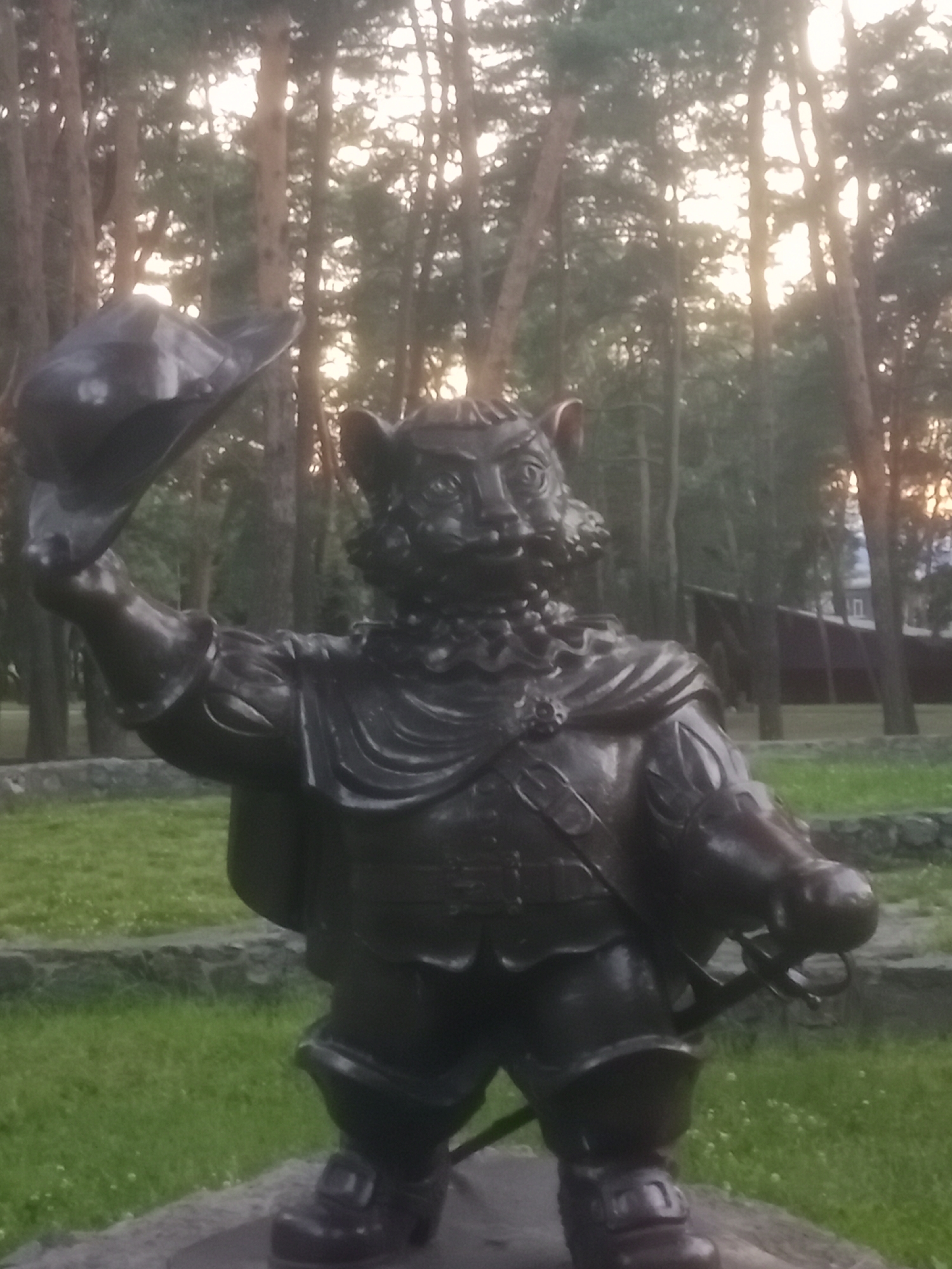 A dark bronze statue of a cat dressed in historical attire stands in a park. The cat is wearing a wide-brimmed hat, a cape, a ruffled collar, and a tunic with a belt. It holds the hat in its left paw and a sword is sheathed at its side. The statue is positioned on a stone base, and in the background, there are tall trees, suggesting a wooded area. The sky appears overcast with sunlight filtering through the trees. This statue is located in Horishni Plavni, Ukraine.