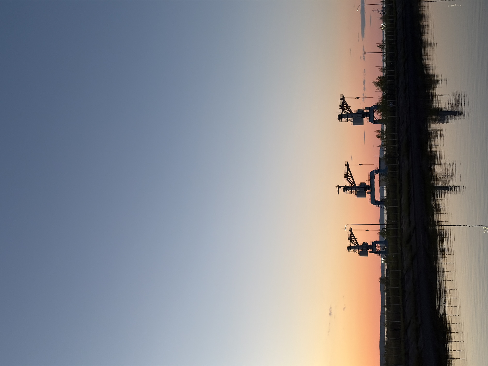 The image depicts a waterfront scene in Chaikovsky, Russia at dusk. Three industrial cranes are silhouetted against the gradient sky, which transitions from blue to orange near the horizon. The cranes are spaced along the shoreline and are reflected in the calm water. Along the shore, there is a row of trees and streetlights, also silhouetted. The water surface shows some light ripple patterns.