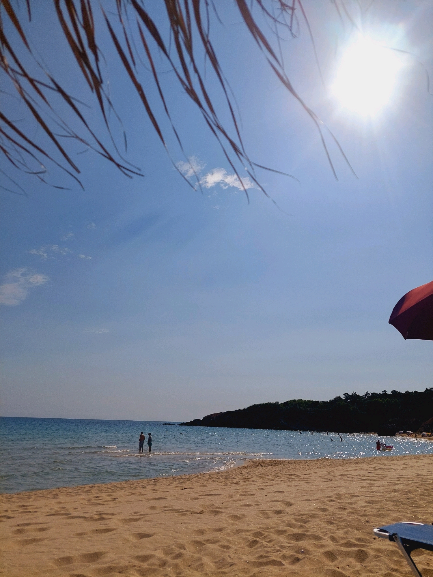 This image depicts a sunny day at a beach, likely in the vicinity of Sozopol, Bulgaria, given the context. In the foreground, a sandy beach stretches towards the shimmering turquoise waters of the sea. The water is calm with small waves gently lapping the shore. Two individuals are standing in the shallow water, facing the sea, with their backs to the camera. Further in the distance, more people can be seen swimming and enjoying the water, with several beach umbrellas dotting the shoreline. A lush green, tree-covered hill forms the backdrop along the coast. The sky is a vibrant blue with a few wispy clouds, and the sun shines brightly, creating a strong lens flare in the upper right portion of the image. Partially obscuring the top of the frame are dry, straw-like fronds, possibly from a beach umbrella or palm tree. A portion of a red umbrella is visible in the far right of the frame. The scene conveys a peaceful and idyllic beach atmosphere.