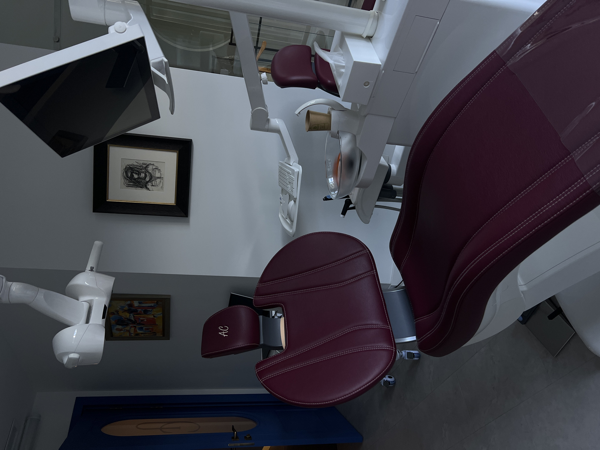 The image shows the interior of a dental office, likely located in Yerevan, Armenia. A modern dental chair with a burgundy leather seat and armrests dominates the scene, positioned at an angle. The chair is equipped with standard dental equipment, including an overhead light and a monitor.
The background features white walls adorned with framed artwork. Below the main chair is a second chair. The lighting appears to be a combination of natural and artificial sources. The floor is tiled in a neutral tone. A door, painted blue with an oval pattern, is visible near the bottom of the frame. Overall, the environment appears clean and professional.